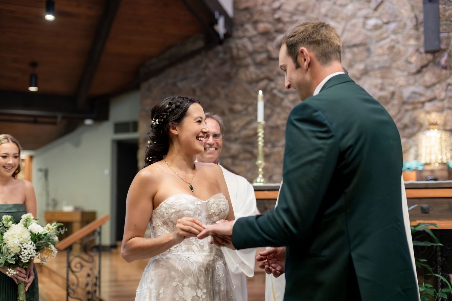 Bride and groom exchange rings at wedding altar, smiling joyfully, officiant behind them in rustic stone chapel.