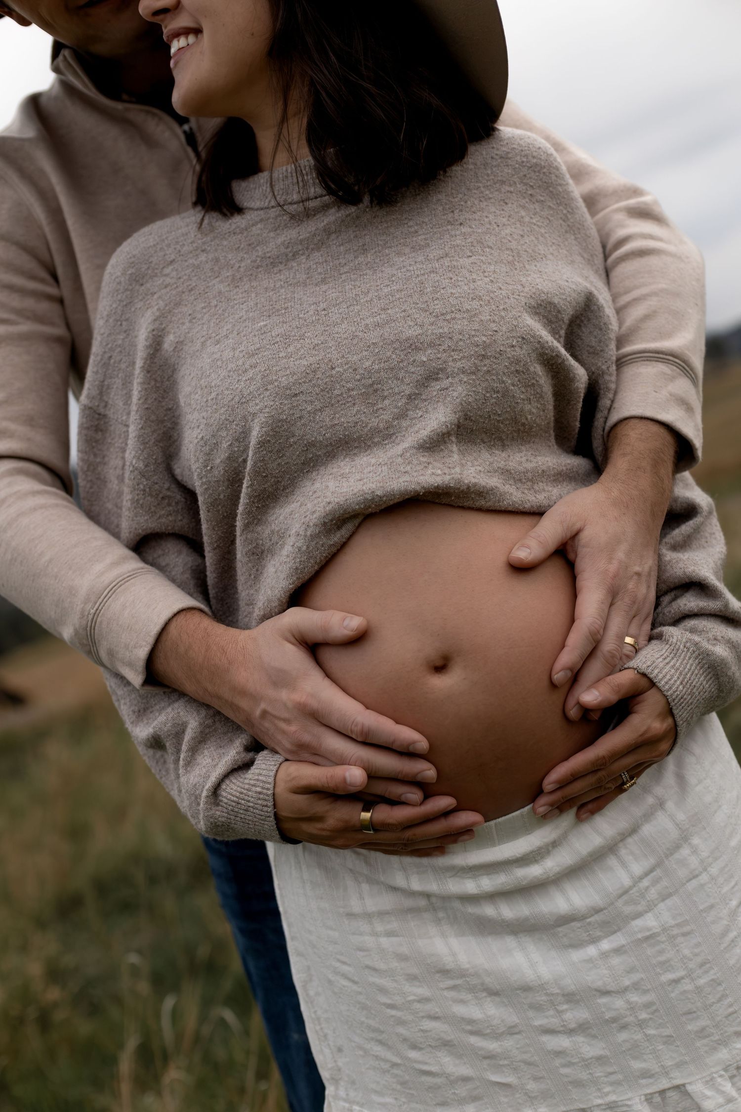 Couple cradling pregnant belly outdoors, wearing neutral tones, sharing a tender moment in a field.