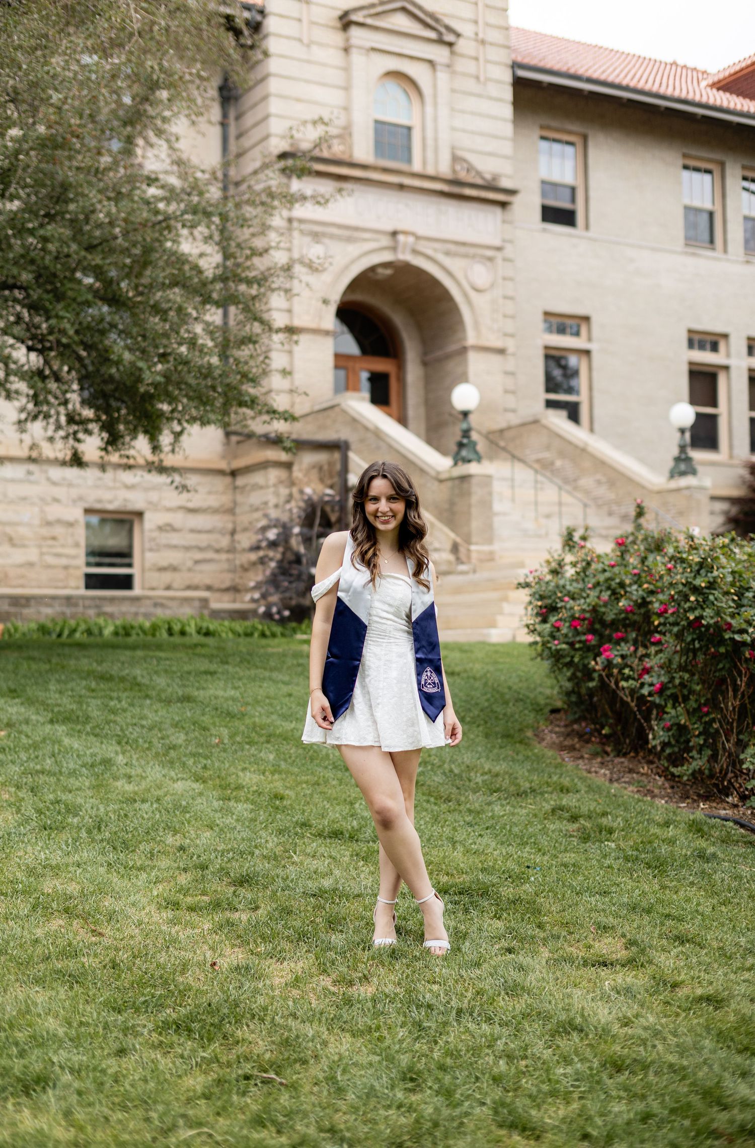 Young woman in white dress and navy blazer poses on green lawn in front of a classic stone university building.
