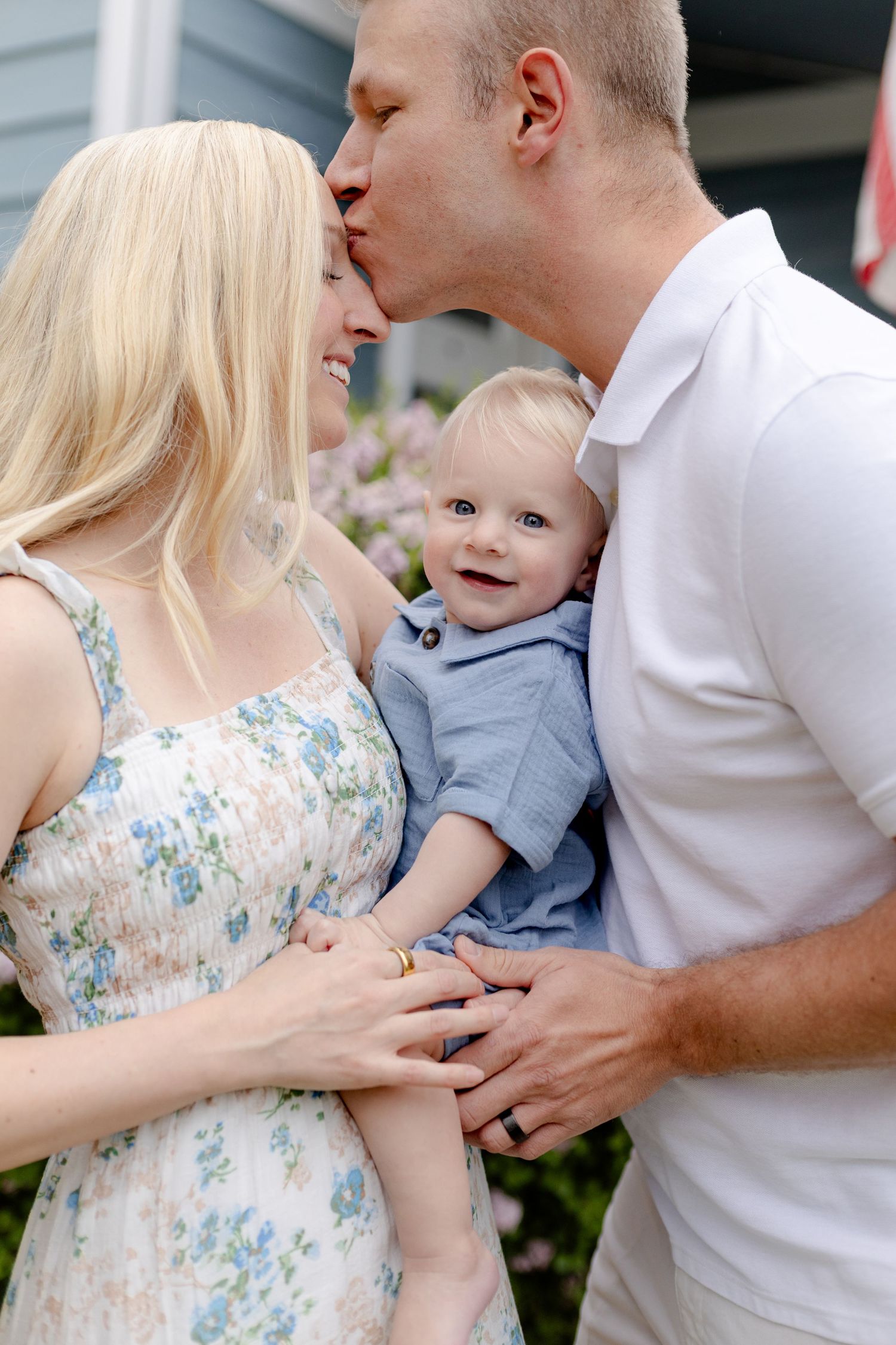 A happy young family with a blonde mother, father kissing her head, and smiling baby in blue outdoors.