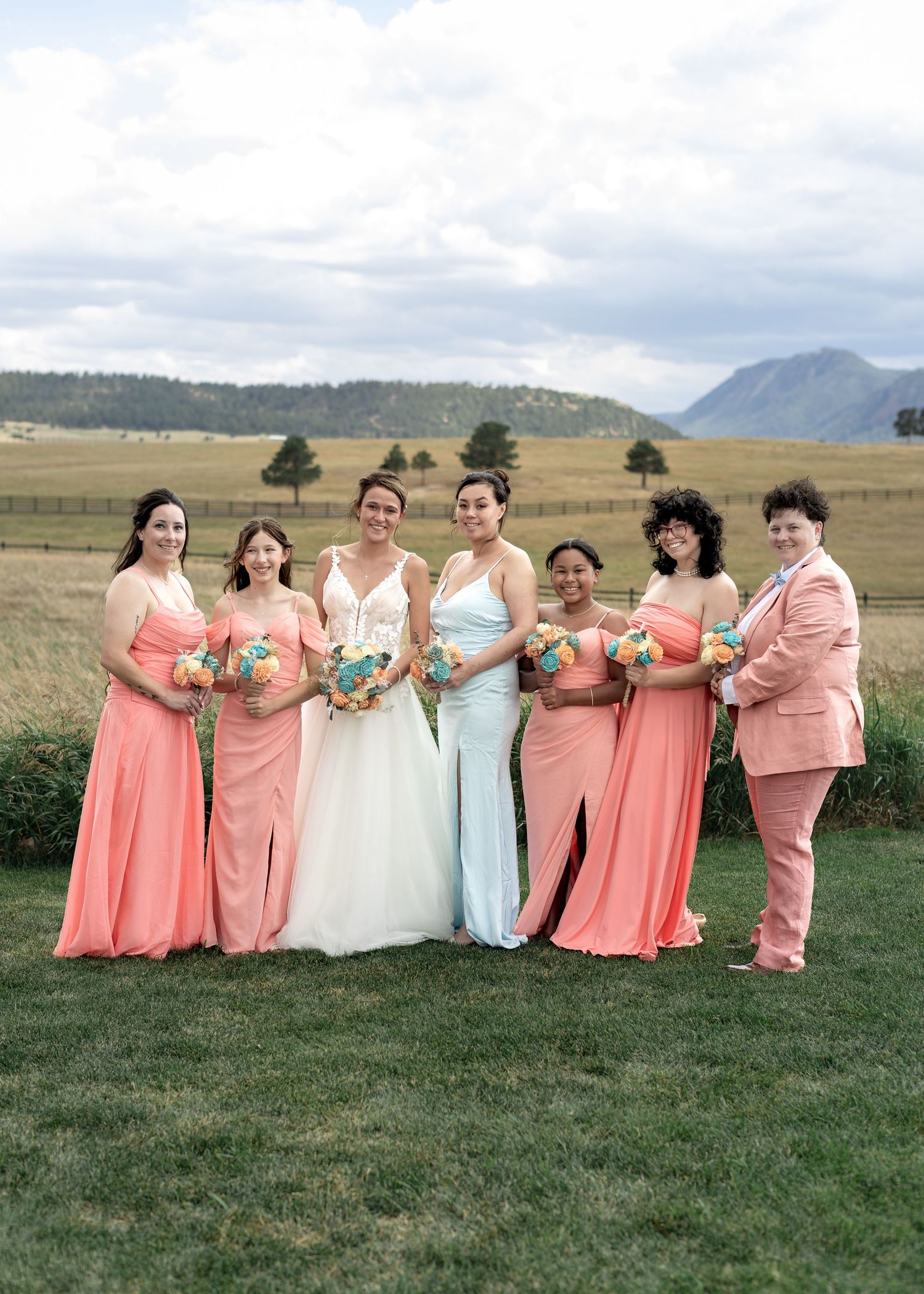Bride poses with five bridesmaids in coral dresses and one in a pink suit at an outdoor mountain wedding venue.