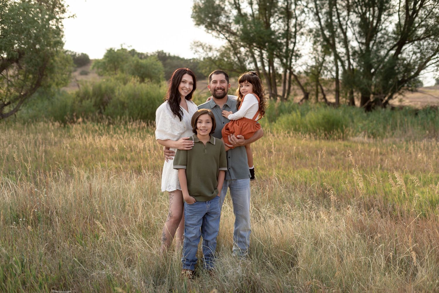 Family of four posing in golden meadow with lush green trees, parents holding toddler during outdoor portrait session.
