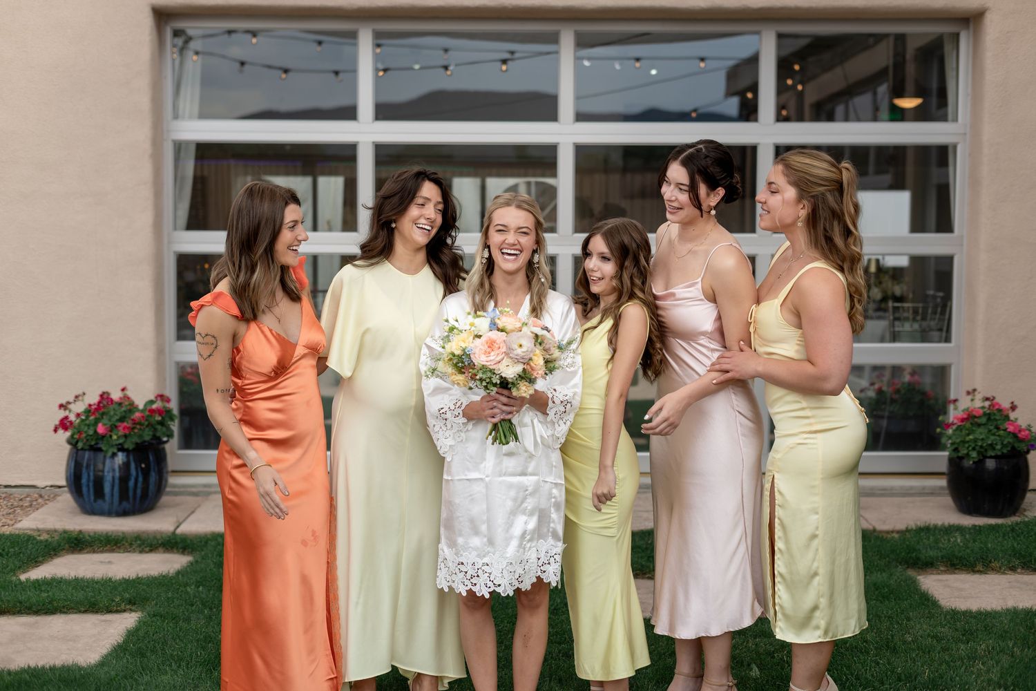 Bride in white dress holds bouquet surrounded by smiling bridesmaids in colorful gowns outdoors.