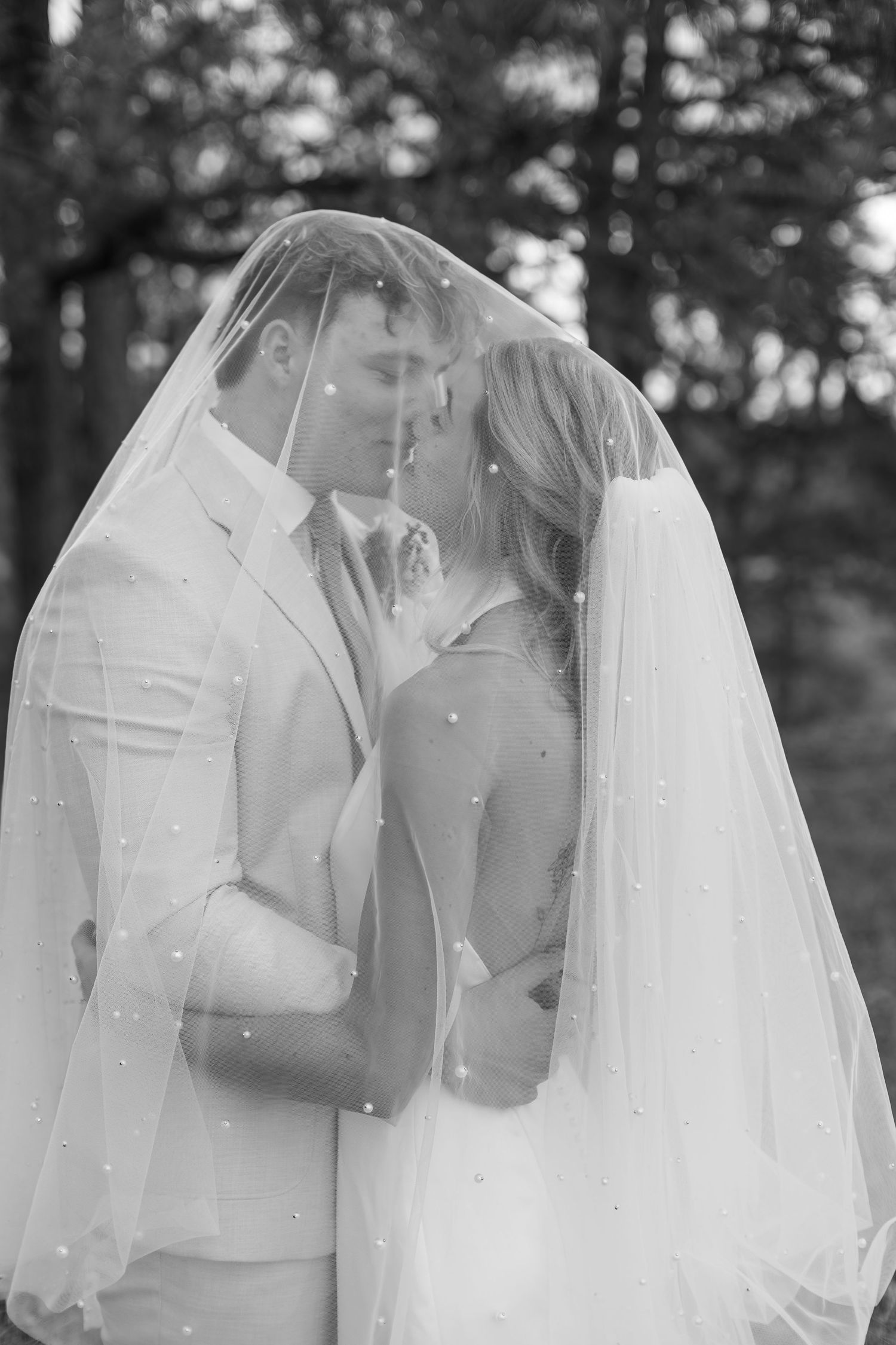 Black and white romantic photo of bride and groom kissing under pearl-studded veil among trees.