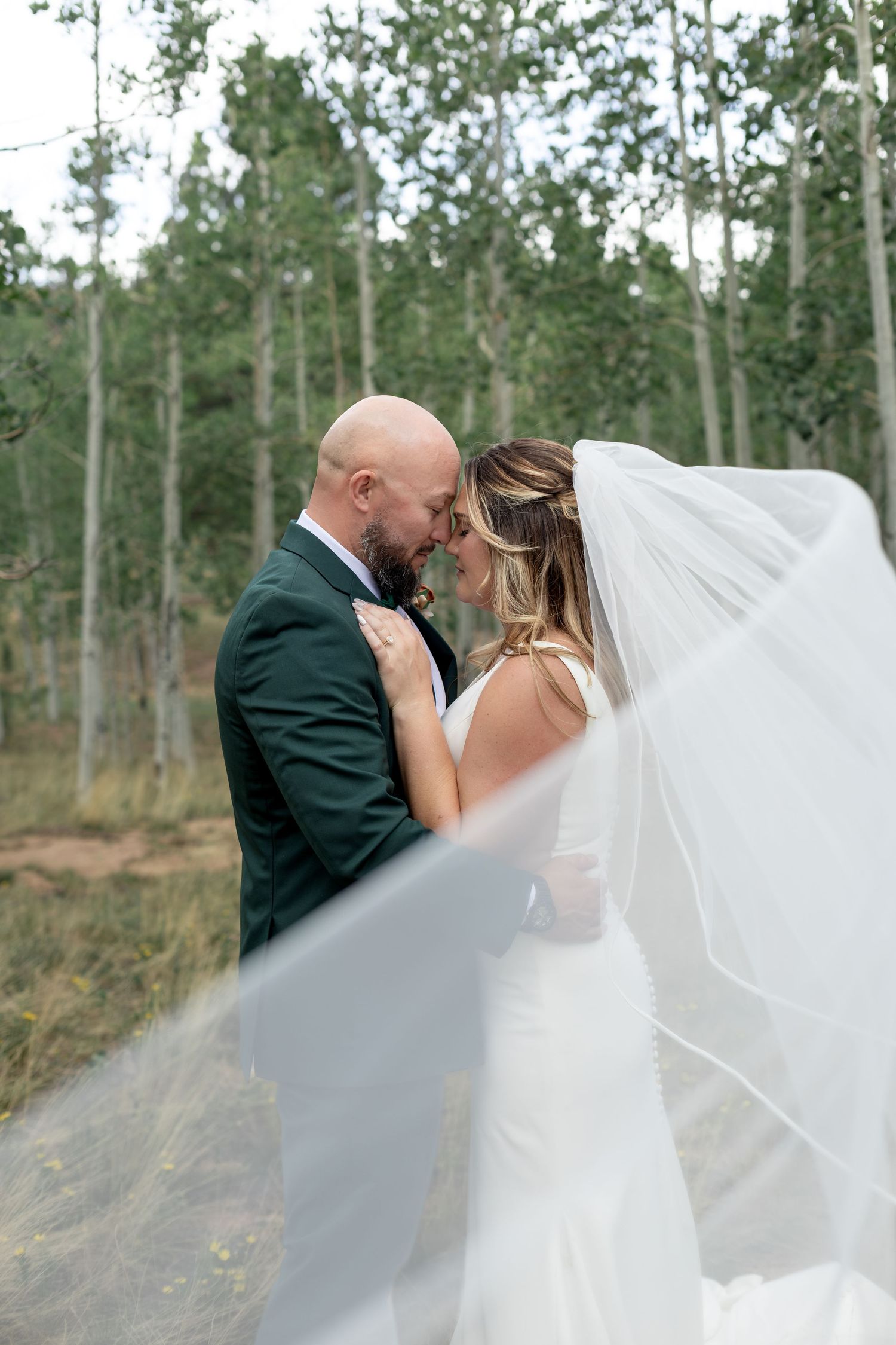 Bride and groom share a romantic forehead touch outdoors, her veil flowing, surrounded by aspen trees.