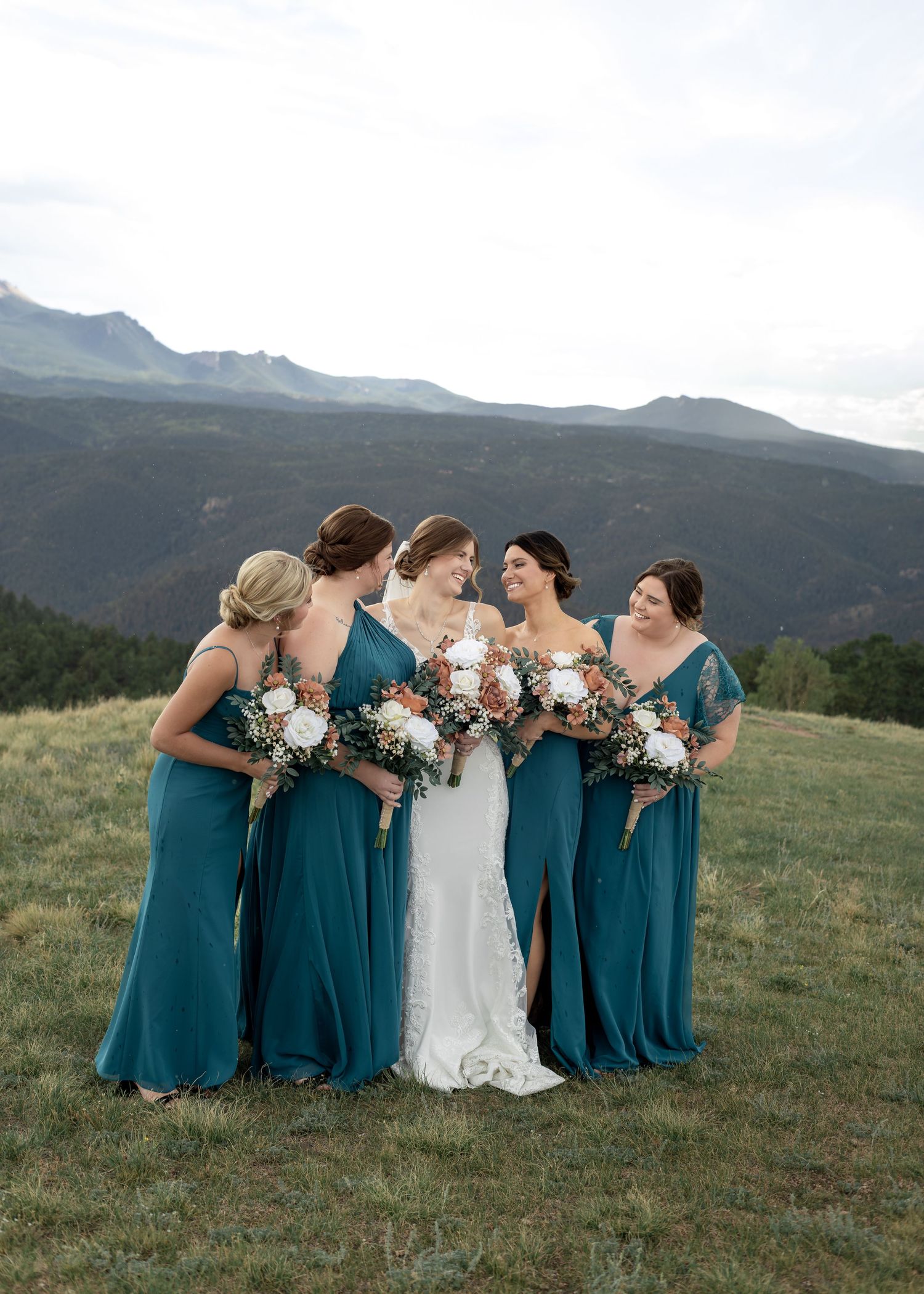 Bridesmaids in teal gowns holding bouquets stand with bride in white dress against mountain backdrop.