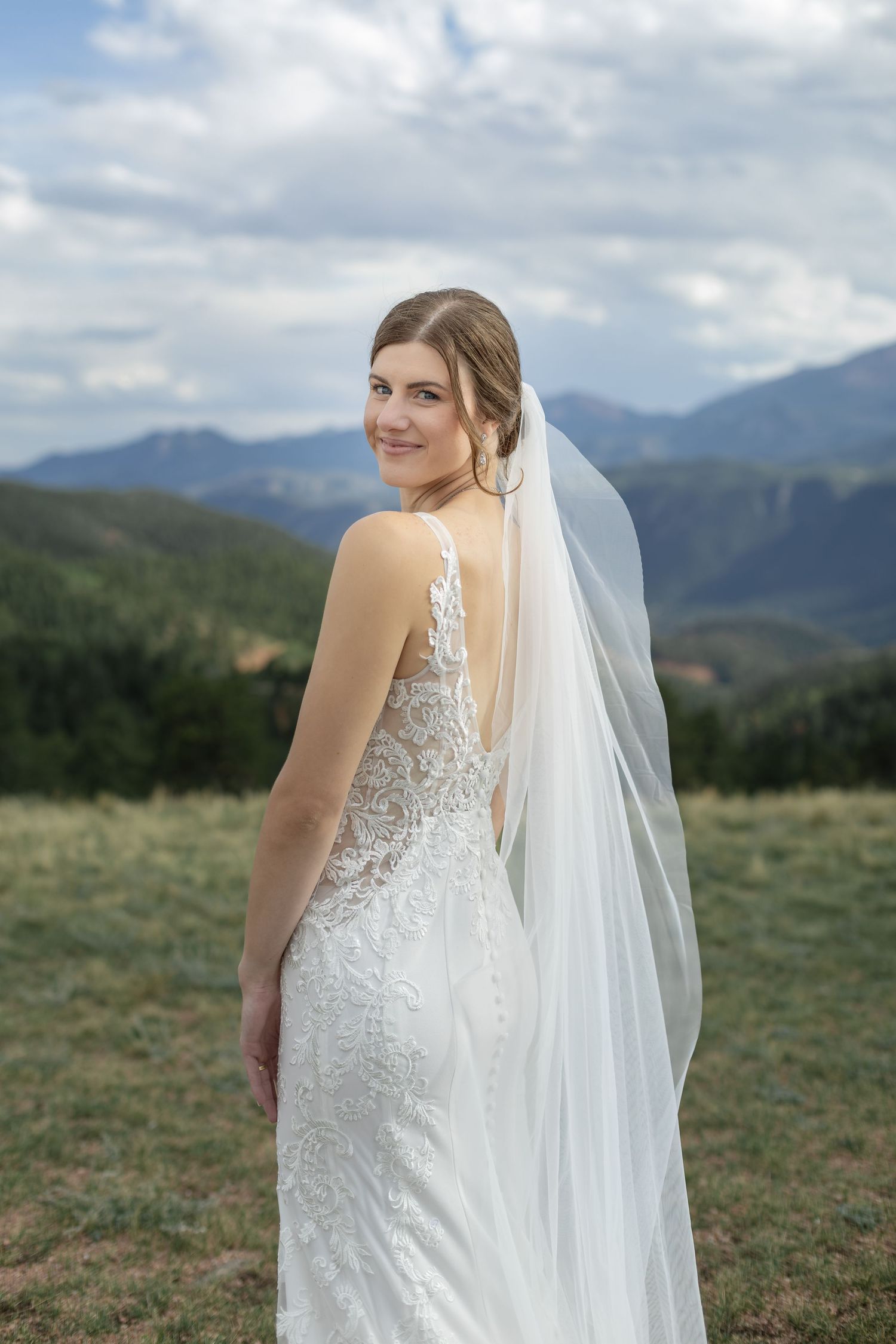 Bride in lace wedding gown and veil poses against scenic mountain backdrop under cloudy sky.