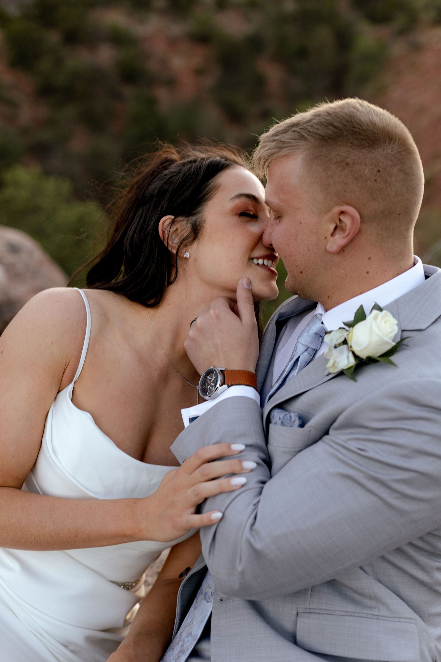 Bride and groom share a romantic moment, nearly kissing, dressed in wedding attire outdoors.