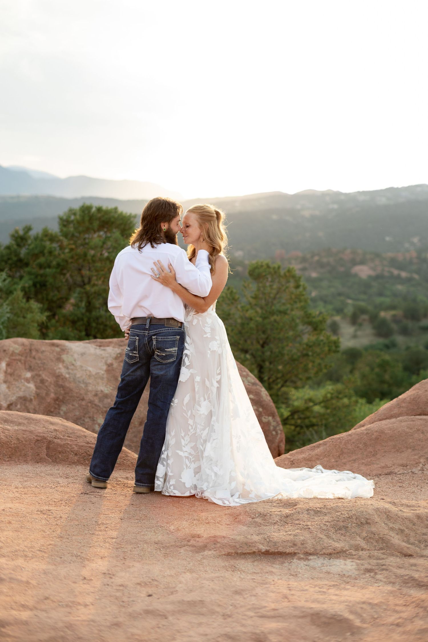Bride and groom share a romantic kiss on red rocks with mountain views at sunset.