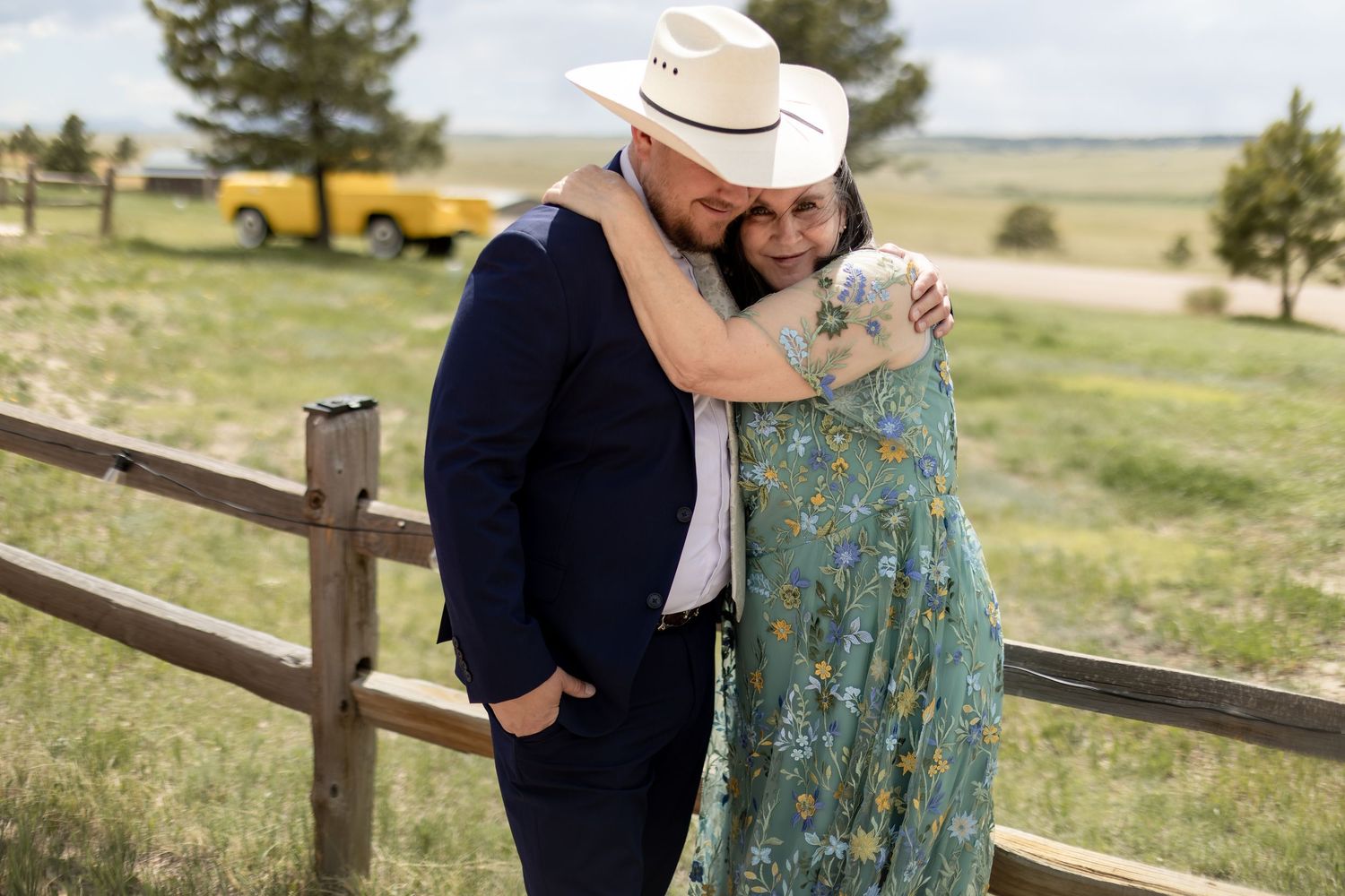 Couple embracing by a wooden fence at a ranch wedding, wearing cowboy hats and formal attire.