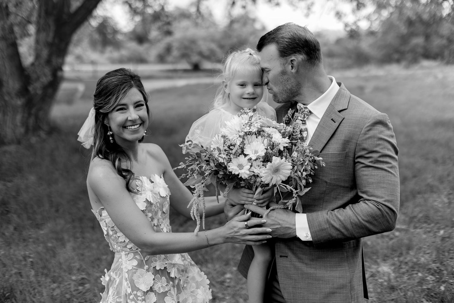 Black and white wedding photo of bride and groom kissing young flower girl holding bouquet outdoors.