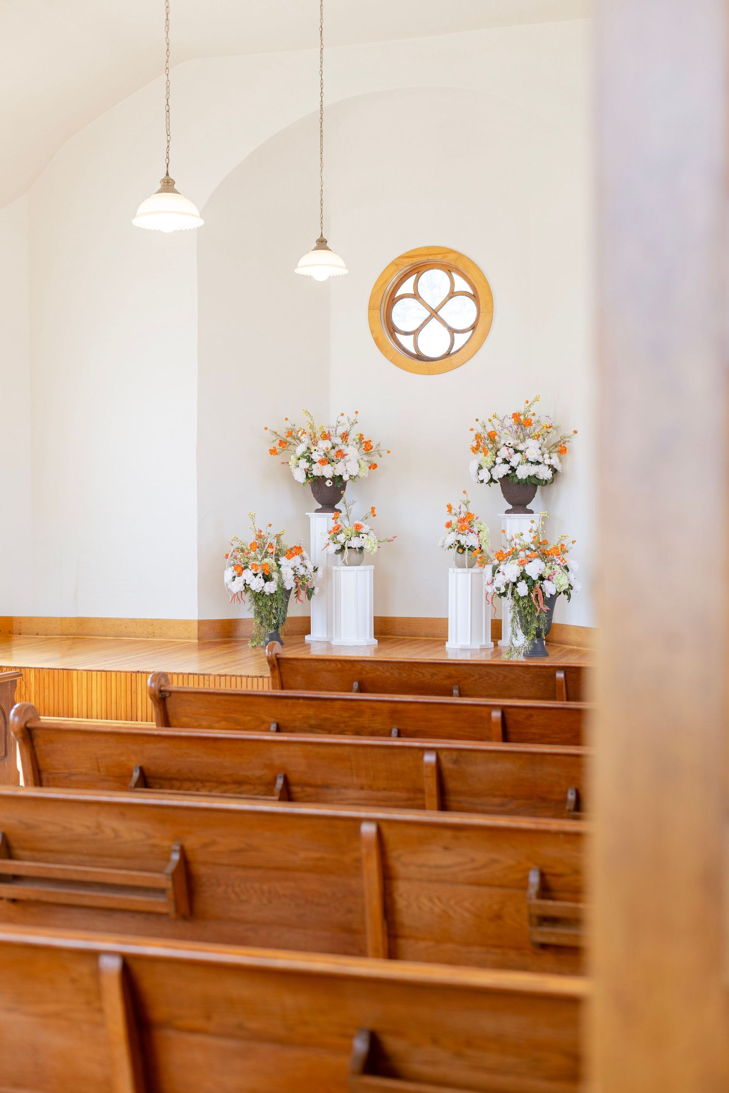 Small chapel interior with wooden pews, white walls, floral arrangements, pendant lights, and a quatrefoil window above the altar.
