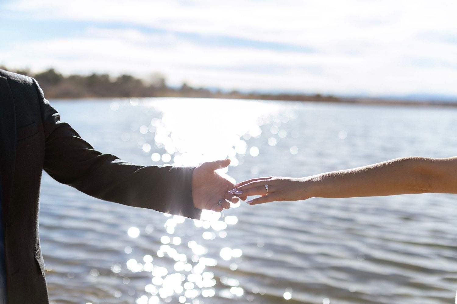 Couple reaching toward each other over a sparkling sunlit lake in an outdoor wedding romantic moment.