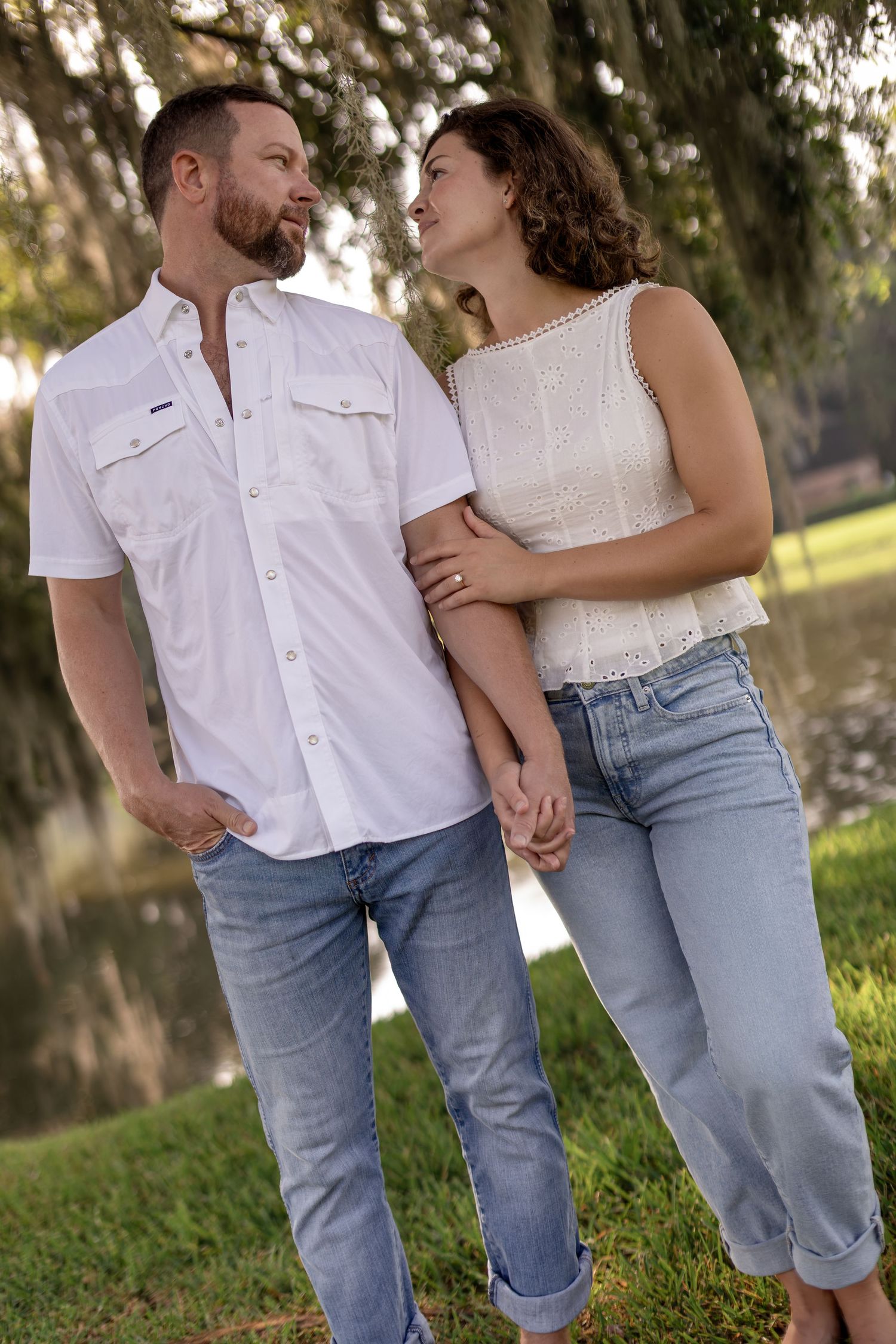 Couple gazes lovingly at each other holding hands near a serene pond surrounded by lush greenery.