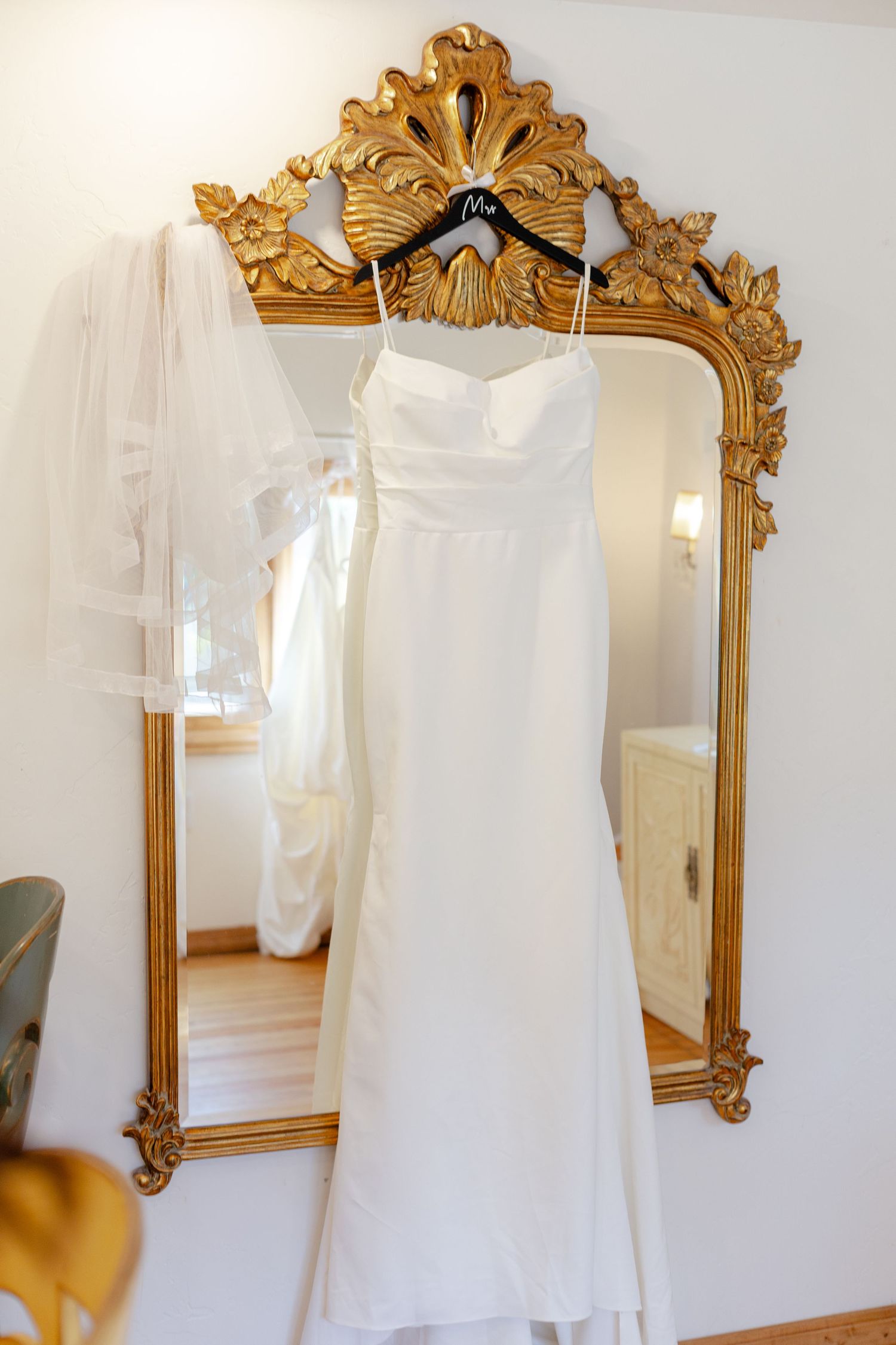 White satin wedding dress hanging on ornate gold mirror with veil beside it in bridal suite.