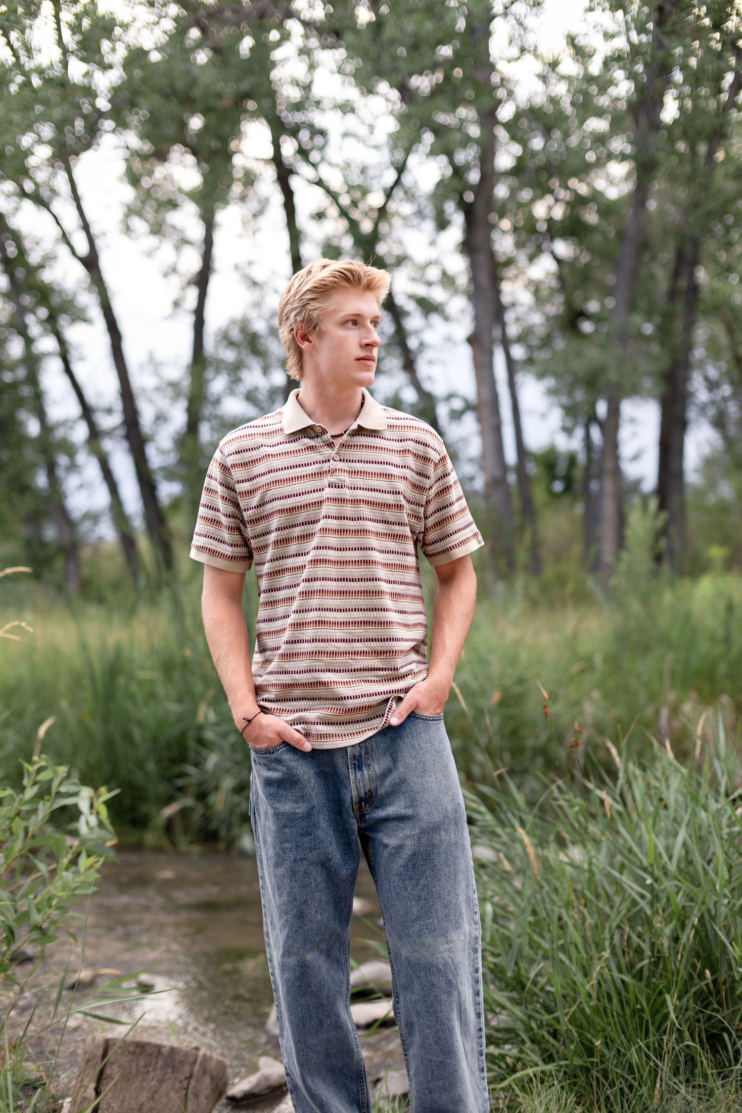 Young man in striped polo shirt and jeans standing outdoors among trees and tall grass.