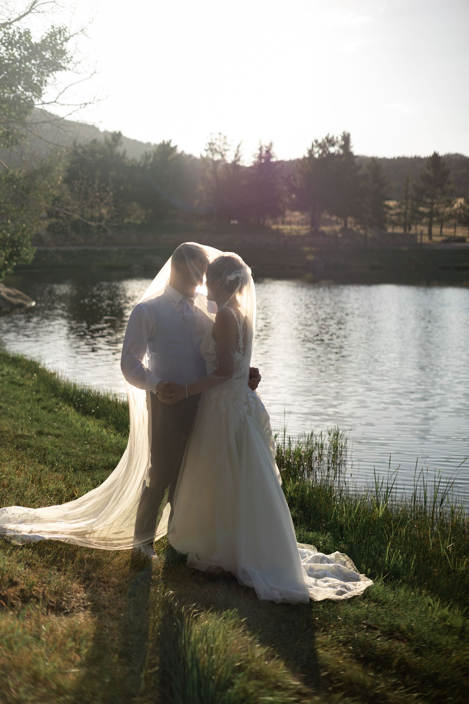 Bride and groom embrace by a sunlit lake, her veil flowing, surrounded by lush greenery and rolling hills.
