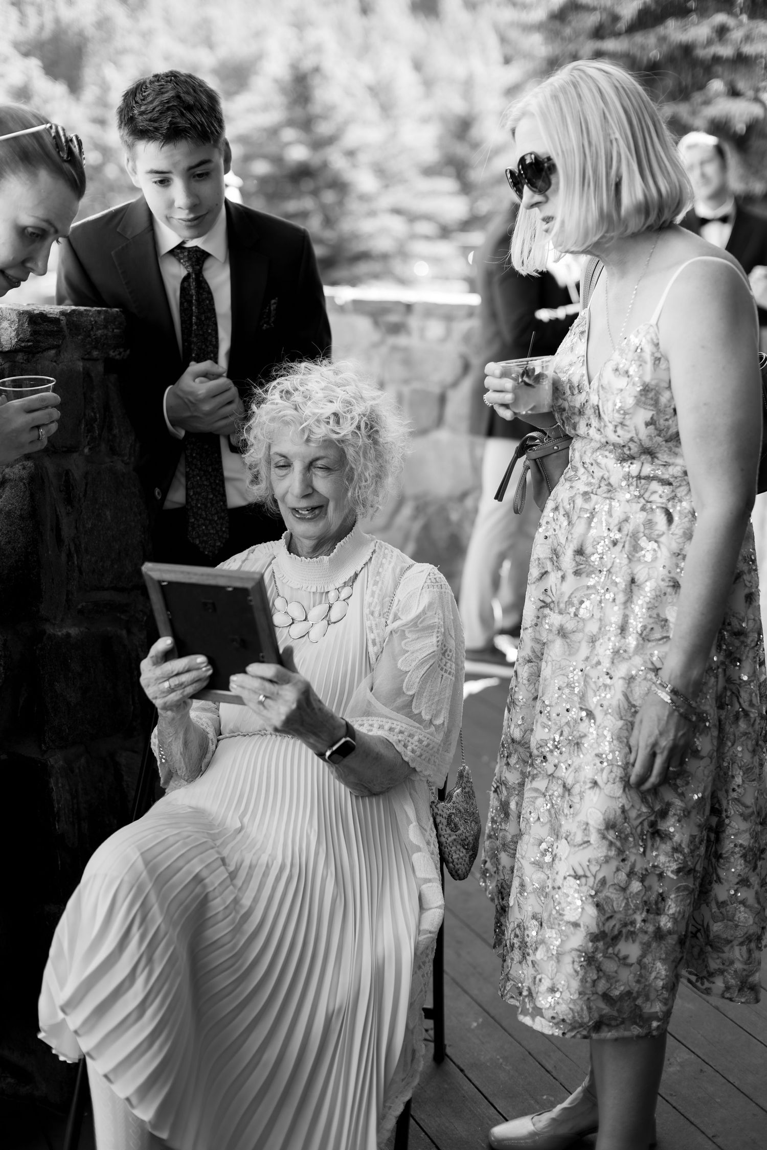 Elderly woman in white dress views tablet at outdoor wedding reception, surrounded by guests in formal attire.