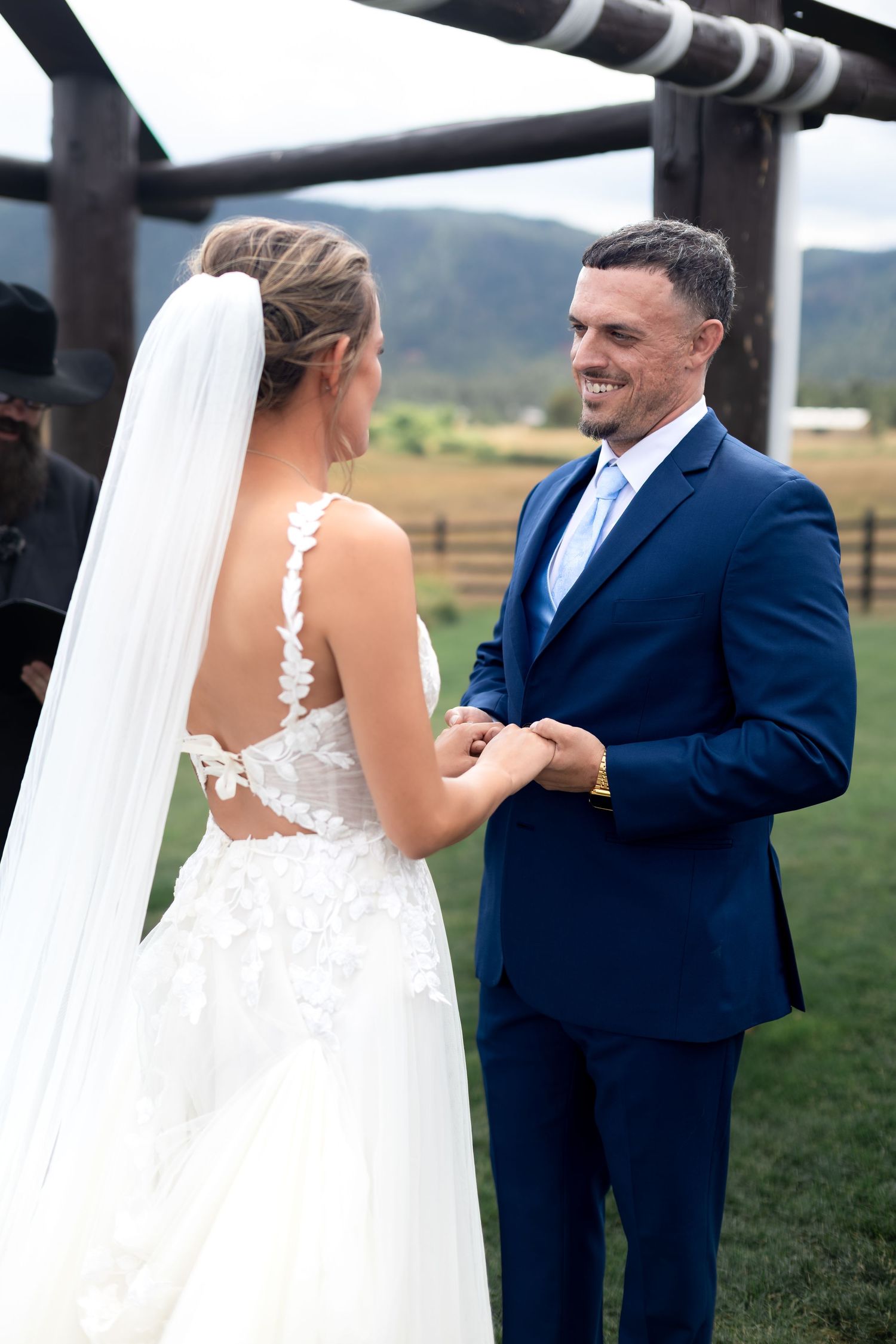 Groom in navy suit smiles at bride in lace backless wedding gown during outdoor ceremony with mountain views.
