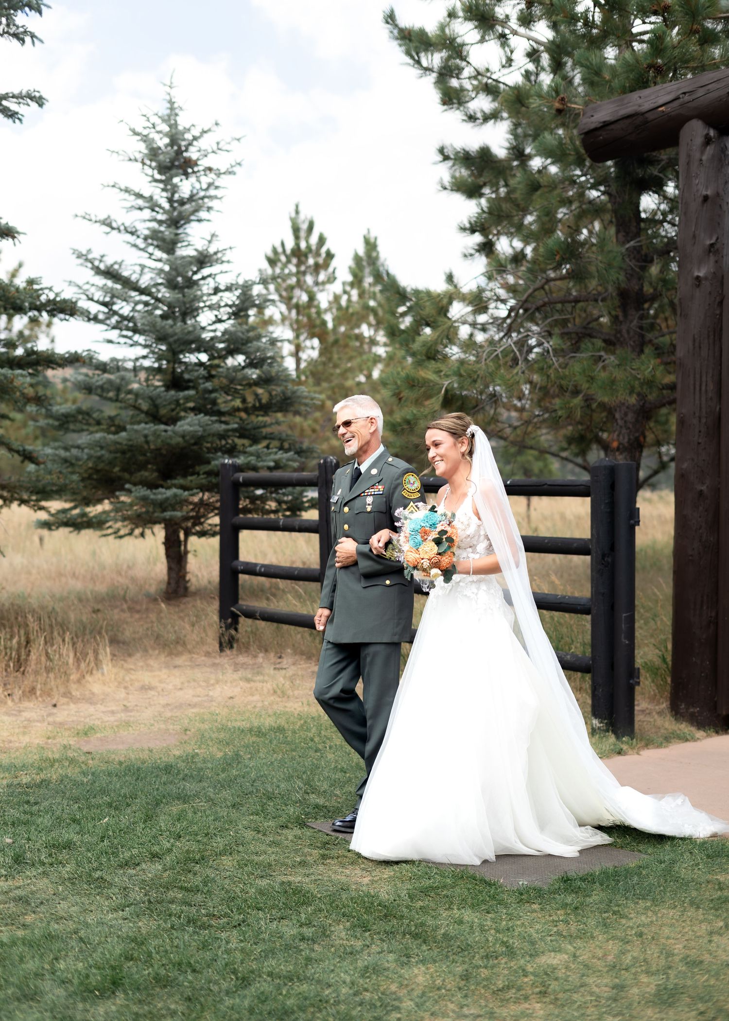 Bride in white gown walks down aisle with father in military uniform at outdoor wedding ceremony.