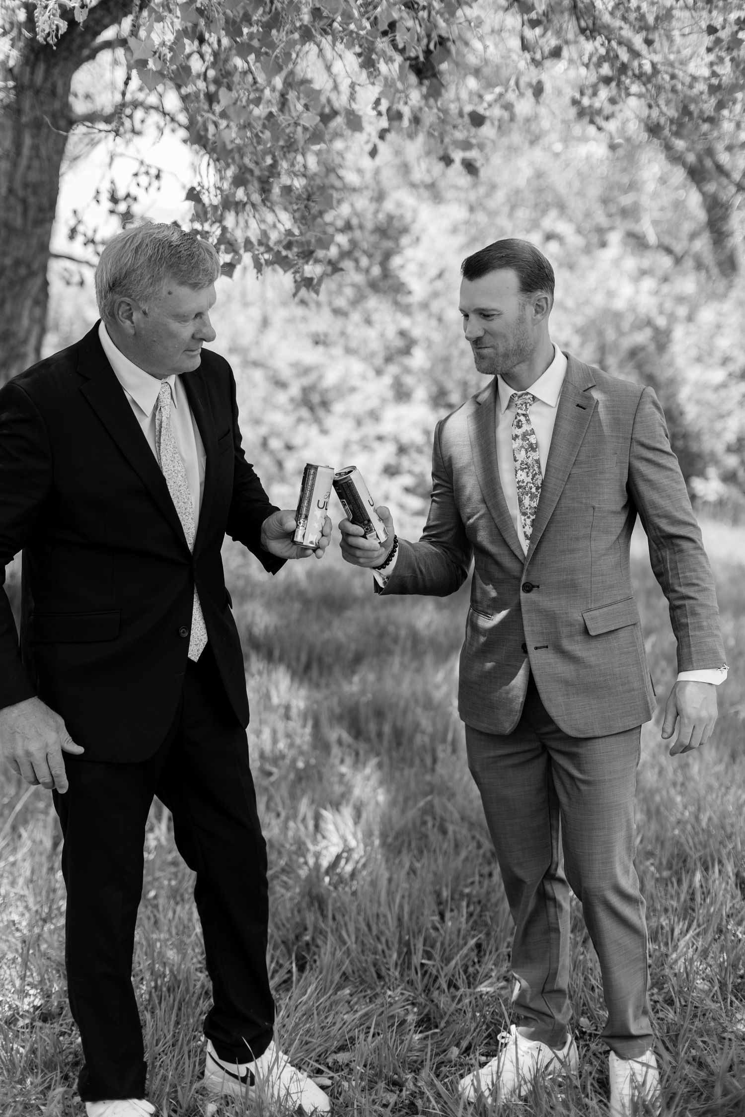 Two men in suits toast with flasks outdoors under trees at a wedding celebration.
