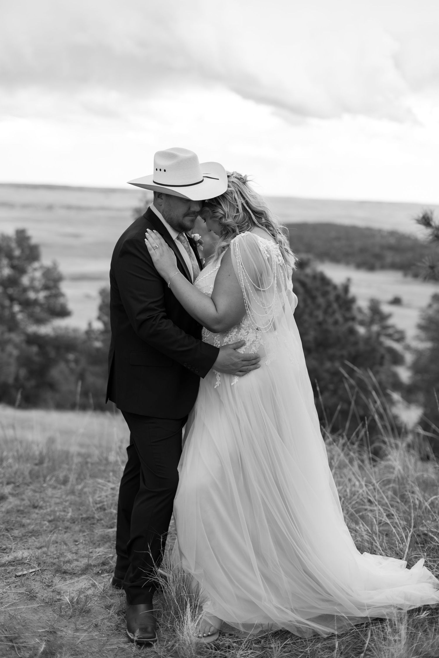 Black and white photo of a bride and groom embracing in a field, groom wearing a cowboy hat, with rolling hills behind.