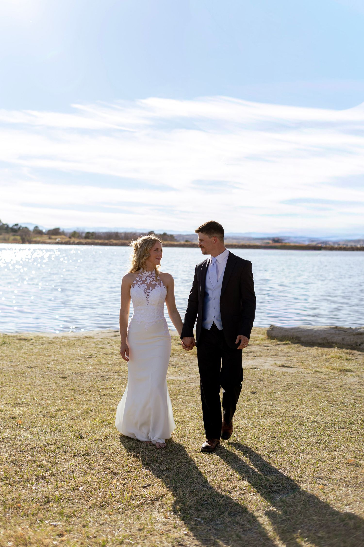 Bride and groom walk hand in hand along a lakeside shore under a bright blue sky on their wedding day.