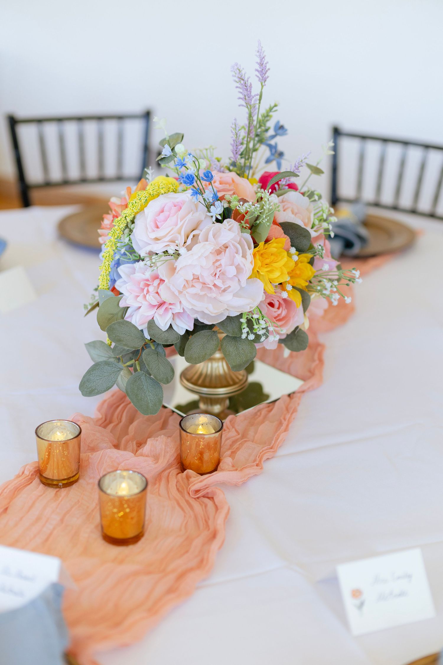 Colorful wedding reception centerpiece with pink peonies, yellow and blue flowers in a gold vase on a peach table runner.