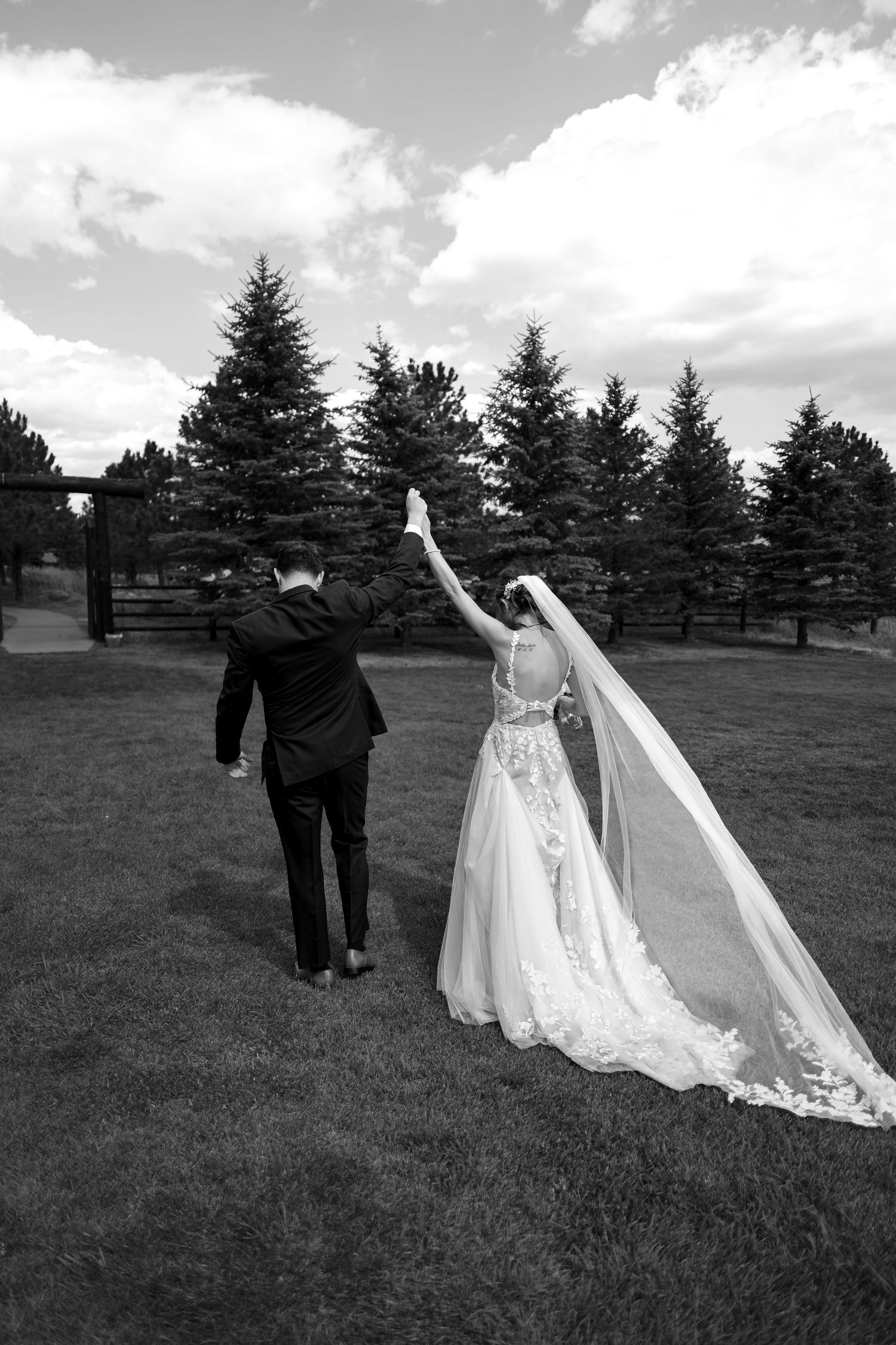 Bride and groom hold hands walking across green lawn with tall pine trees in black and white.