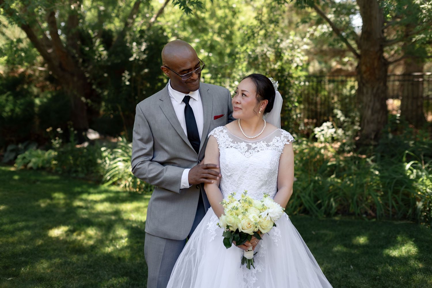 Interracial couple in wedding attire posing outdoors, groom in gray suit, bride in white gown holding bouquet.