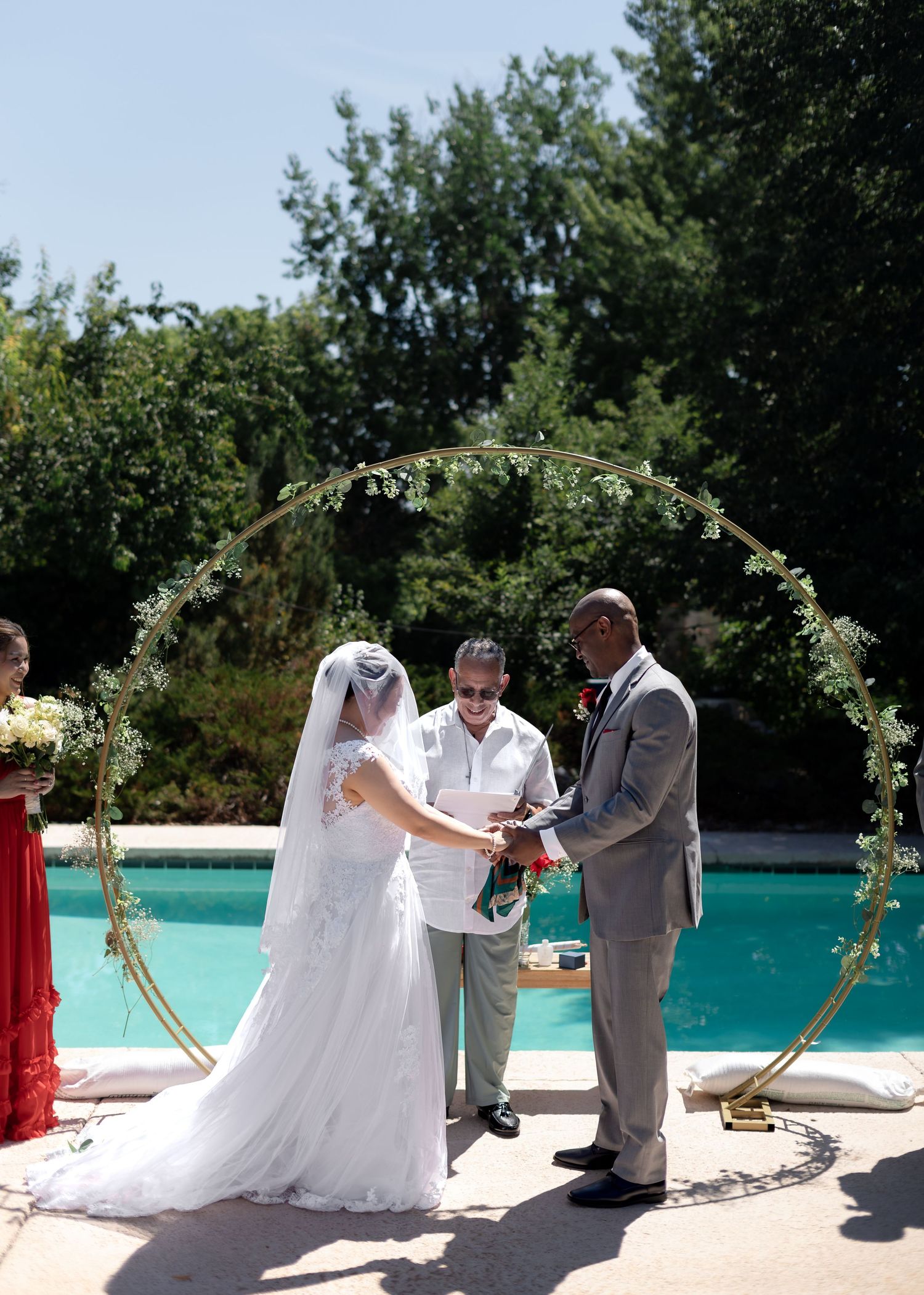 Bride and groom exchange vows at outdoor wedding ceremony by pool under circular floral arch on sunny day.