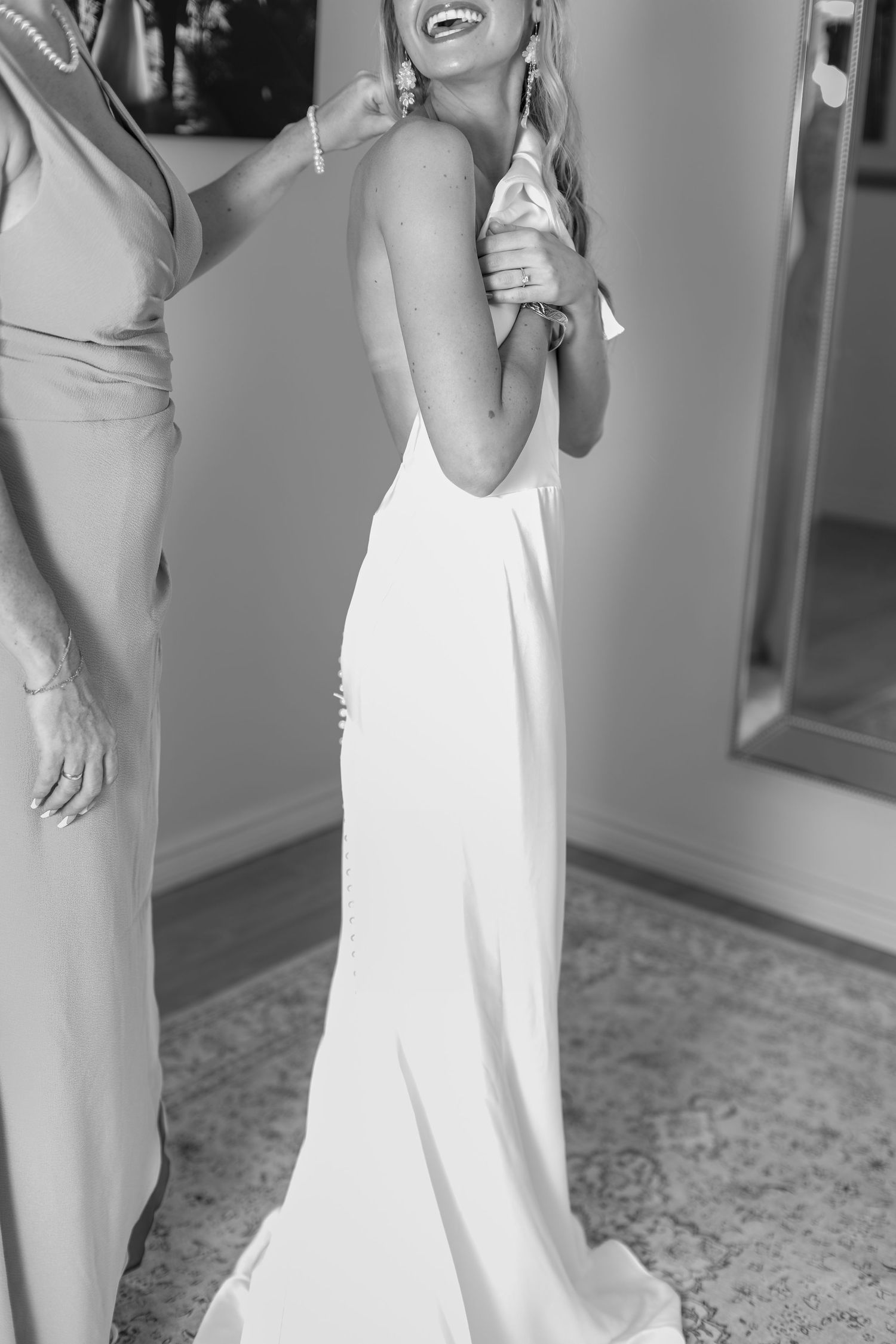 Black and white photo of bride in backless wedding gown laughing as someone helps fasten her earring.