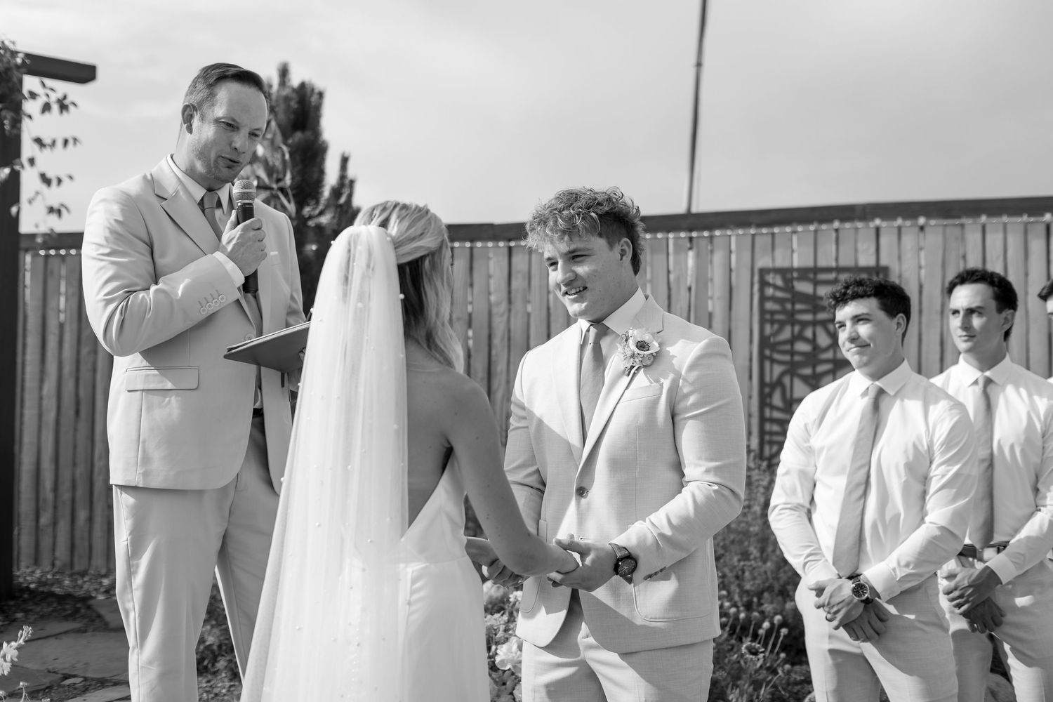 Groom smiles at bride during outdoor wedding ceremony with groomsmen standing nearby in black and white.
