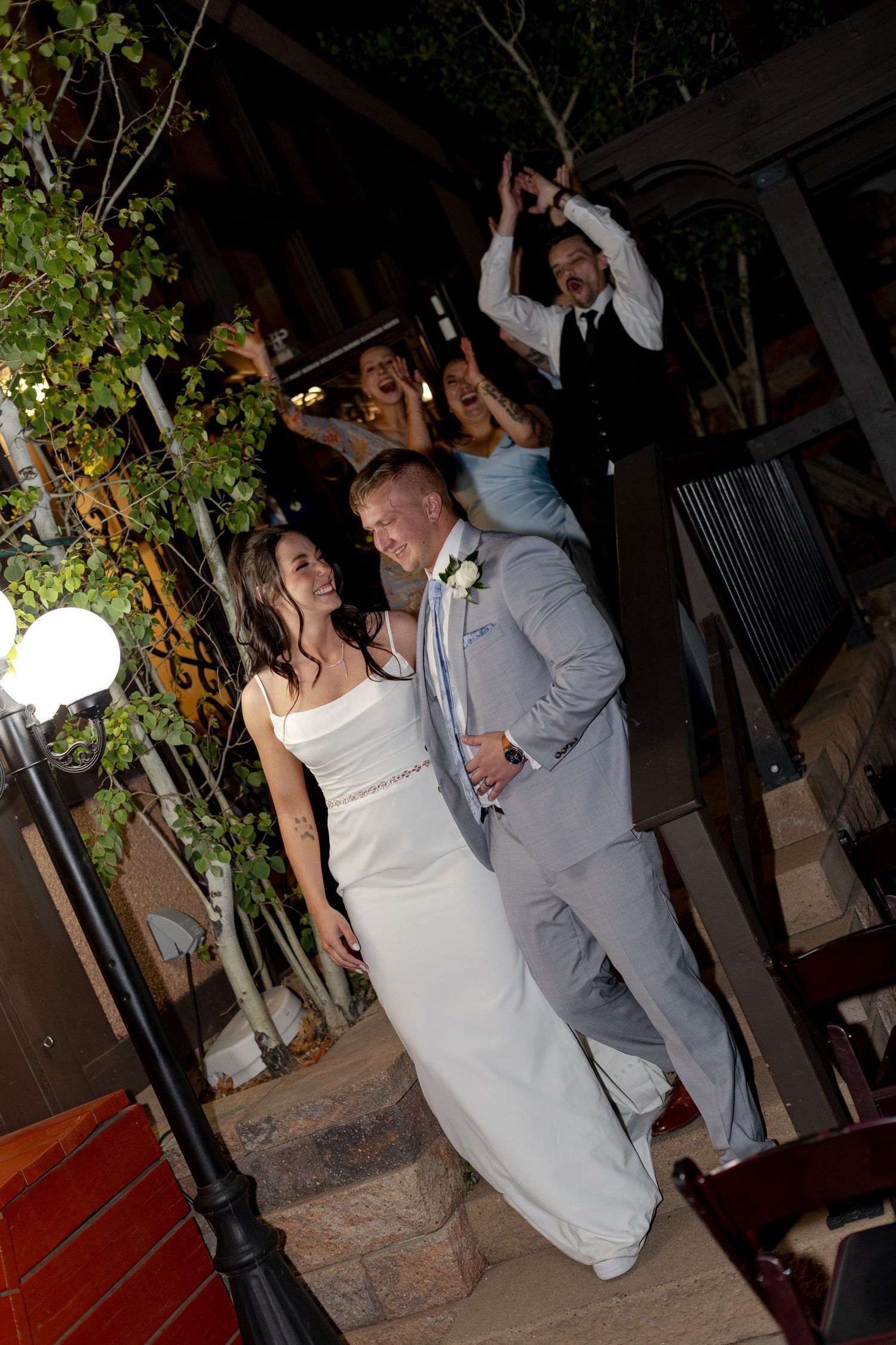 Newlyweds in white gown and gray suit celebrate grand exit with cheering wedding party at night.