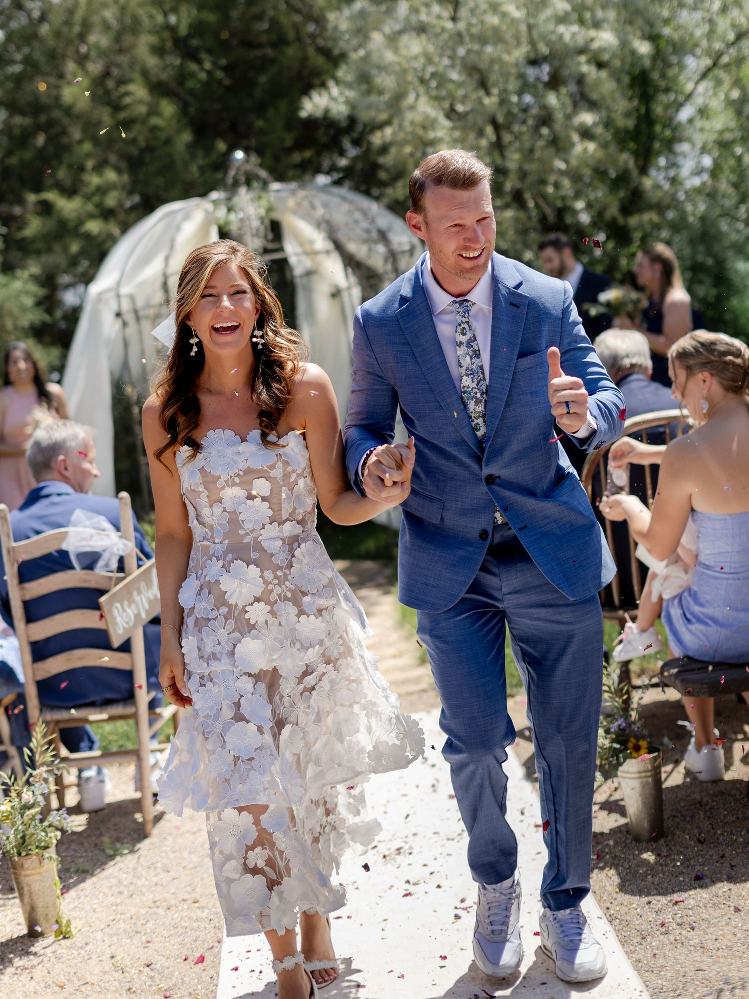 Joyful newlyweds walk down outdoor aisle, bride in floral white dress, groom in blue suit, guests tossing confetti.