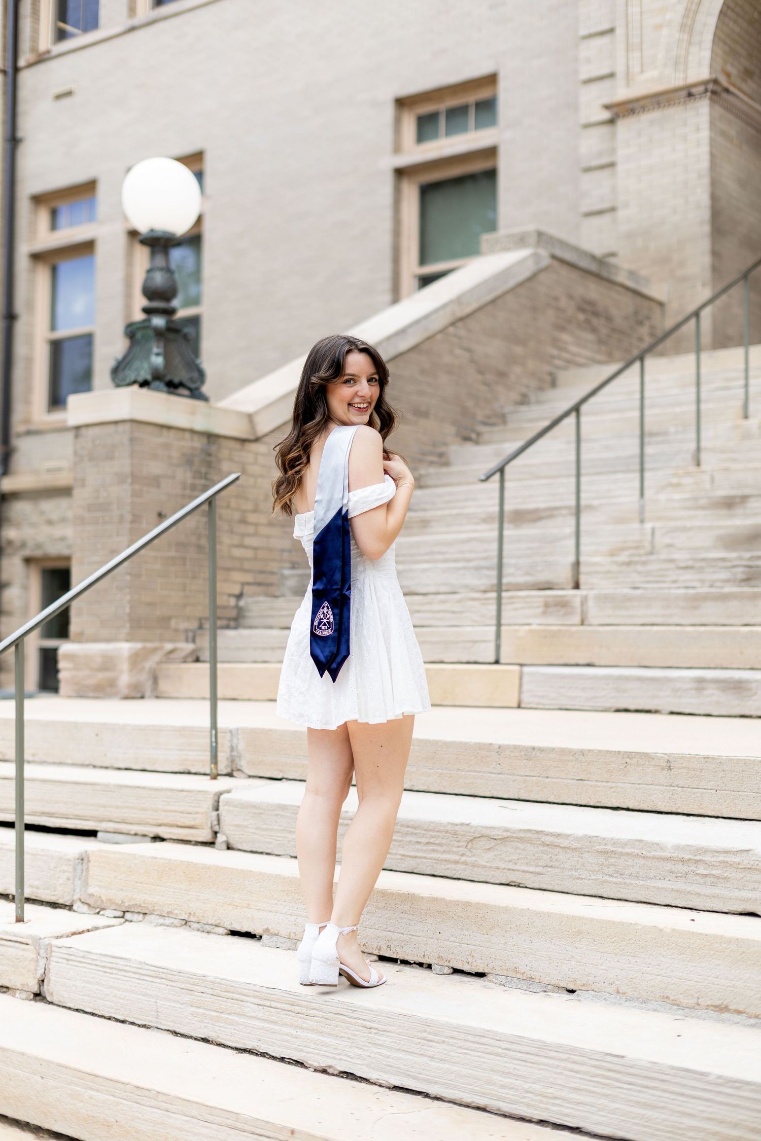 Young woman in white dress with navy graduation stole poses on university steps smiling over shoulder.