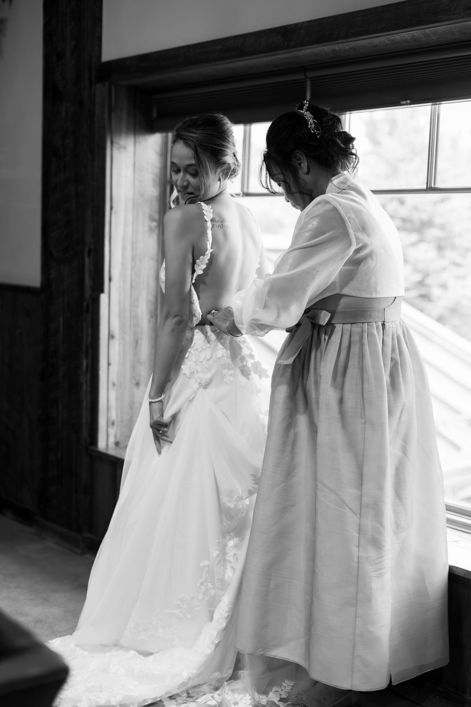 Black and white photo of a bride in a lace gown being helped into her dress by a bridesmaid near a window.