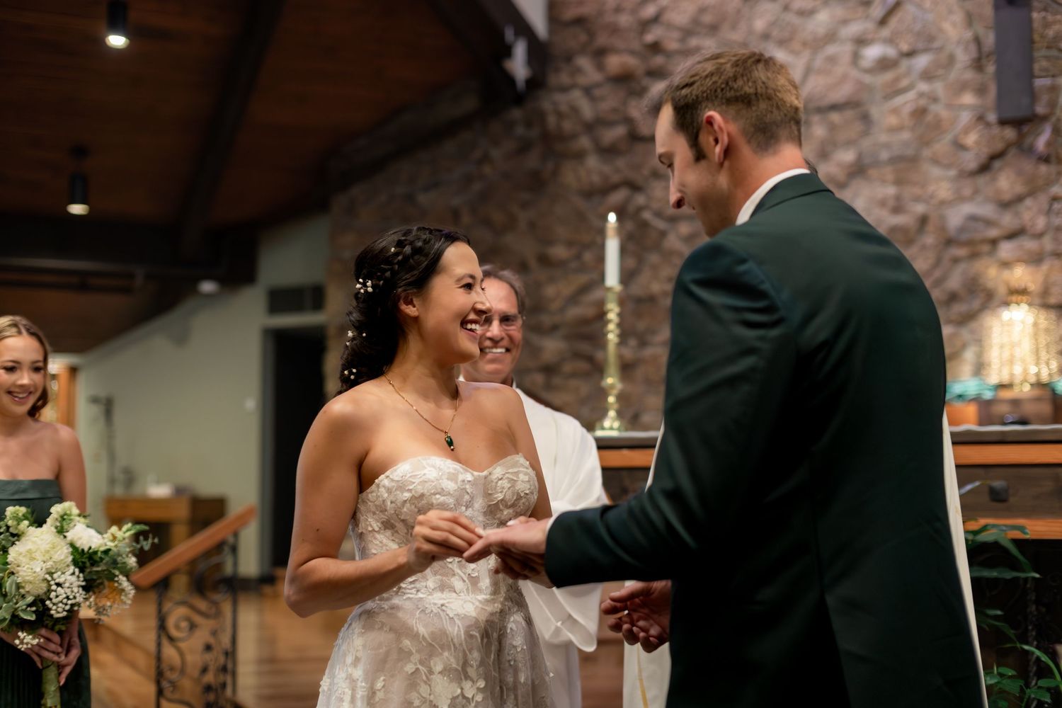 Bride and groom exchange rings at wedding ceremony in rustic stone chapel with officiant smiling behind them.