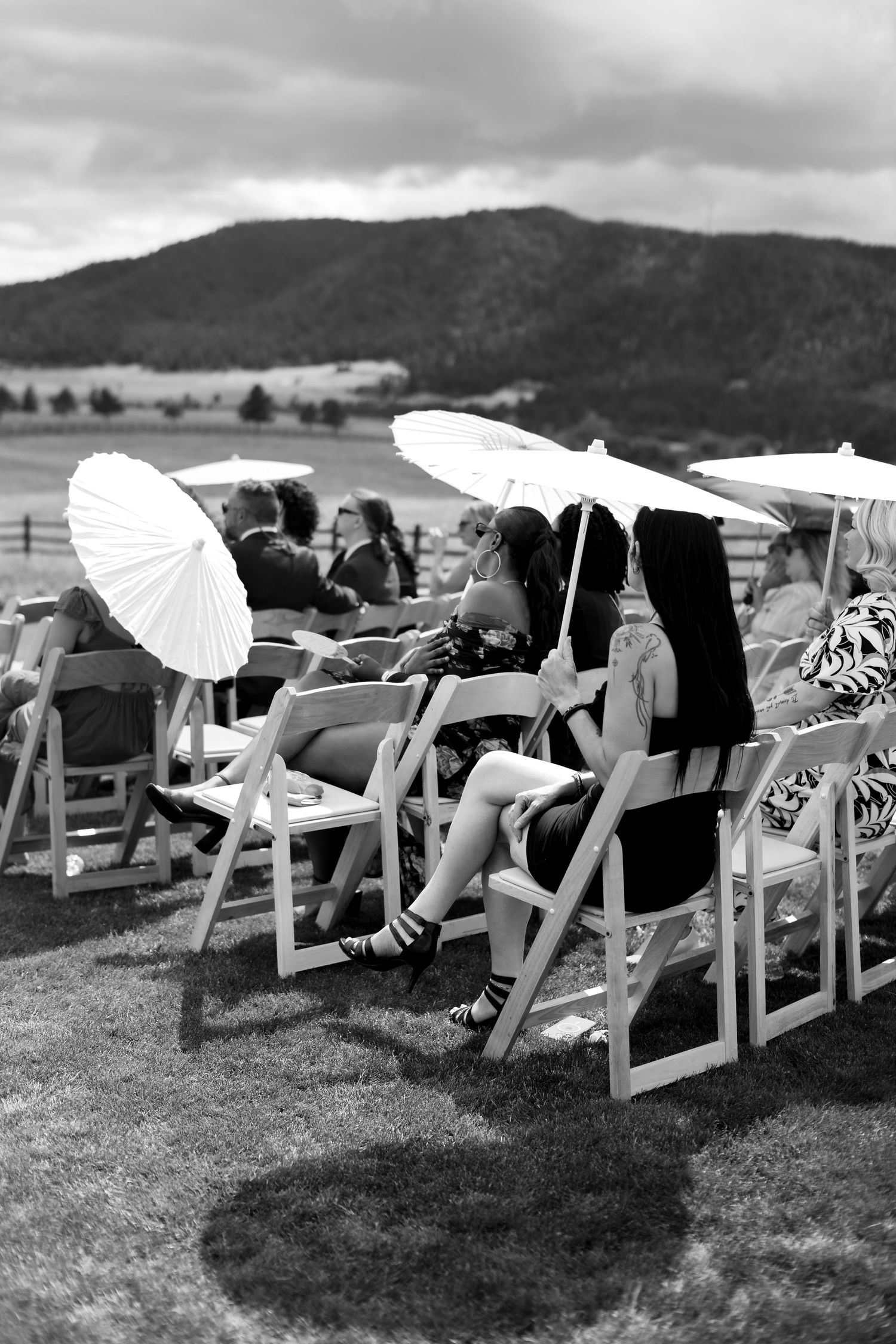 Black and white photo of wedding guests seated outdoors holding white parasols with mountain landscape backdrop.