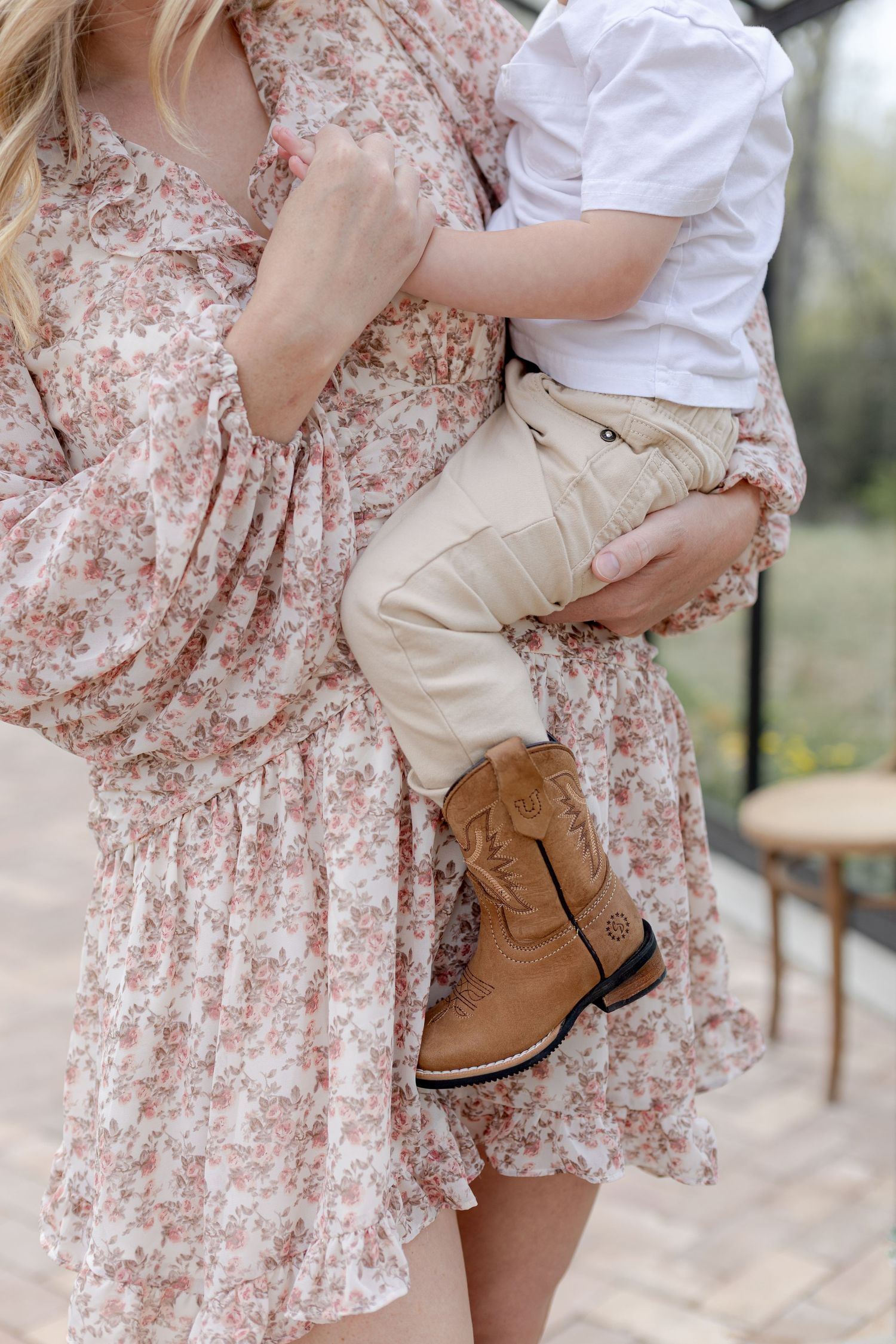 A mother in a floral dress holds a toddler wearing tan pants and brown cowboy boots outdoors.