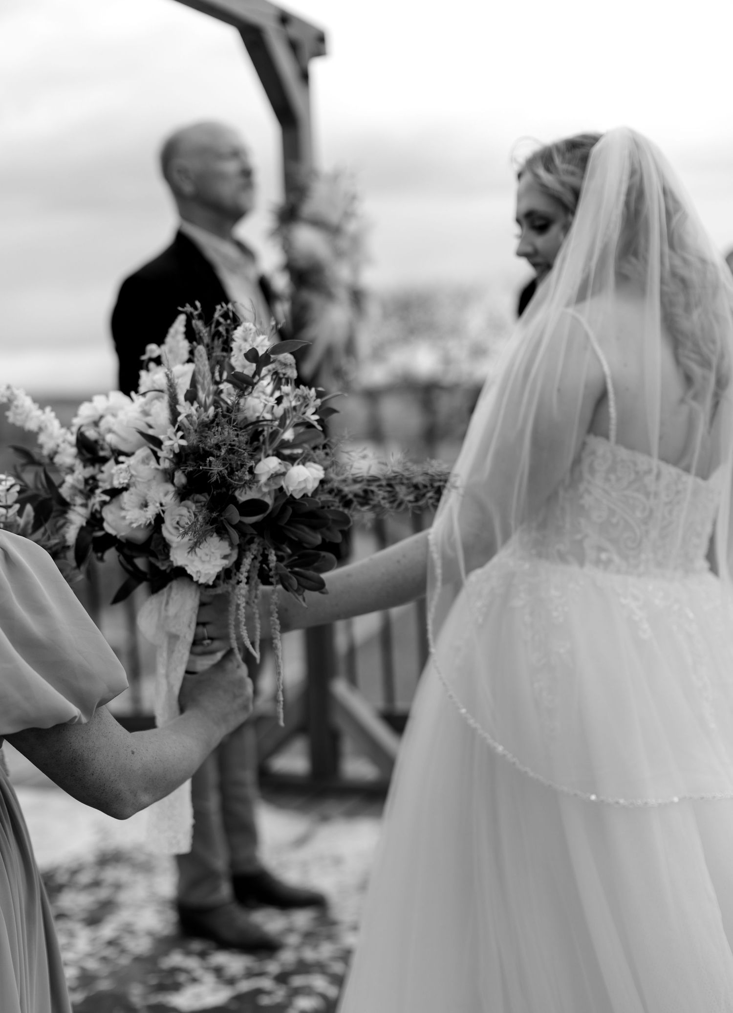 Black and white photo of bride receiving bouquet from bridesmaid at outdoor wedding ceremony.