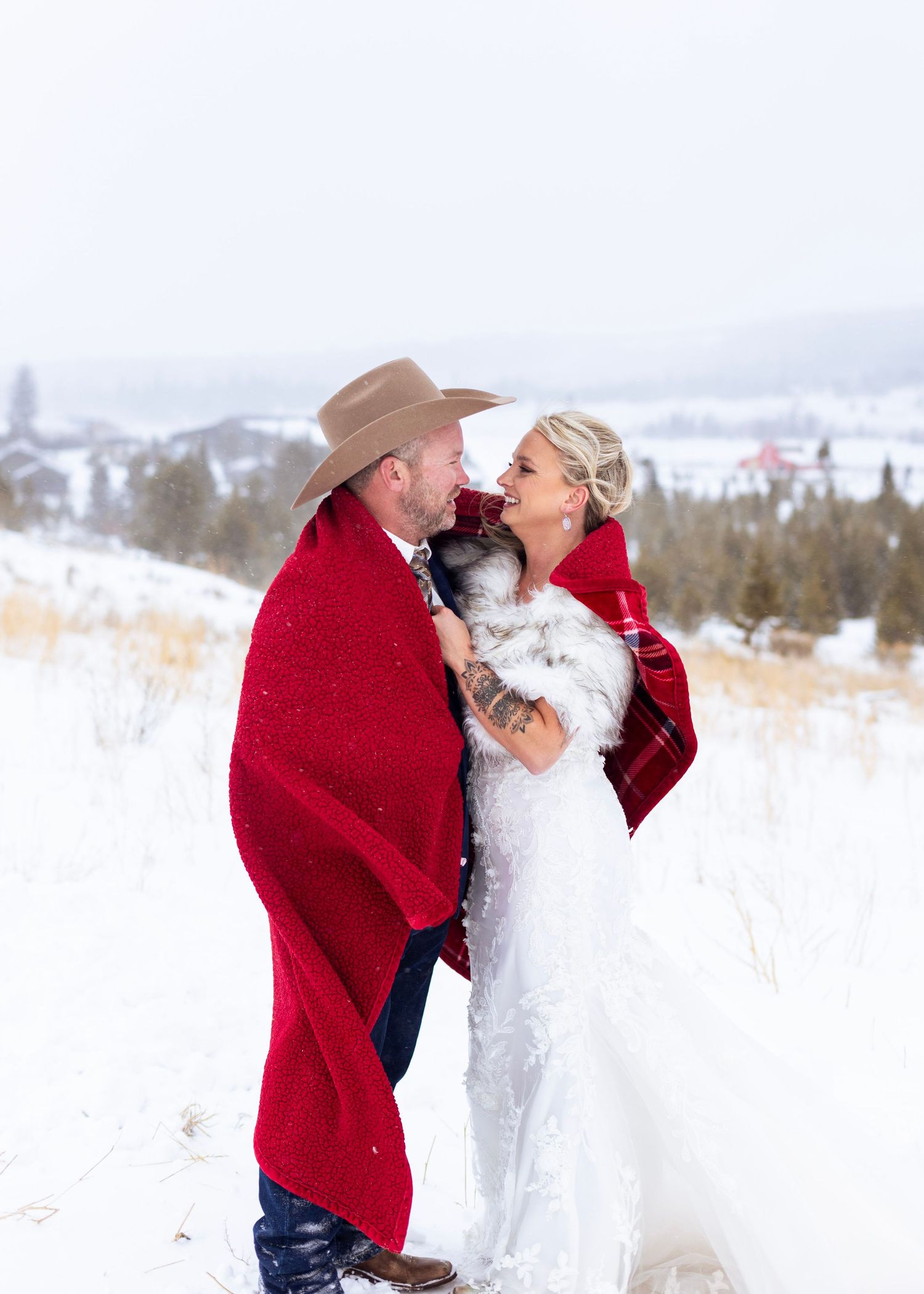 A couple wrapped in a red blanket share a romantic moment in a snowy winter landscape with mountains in the background.