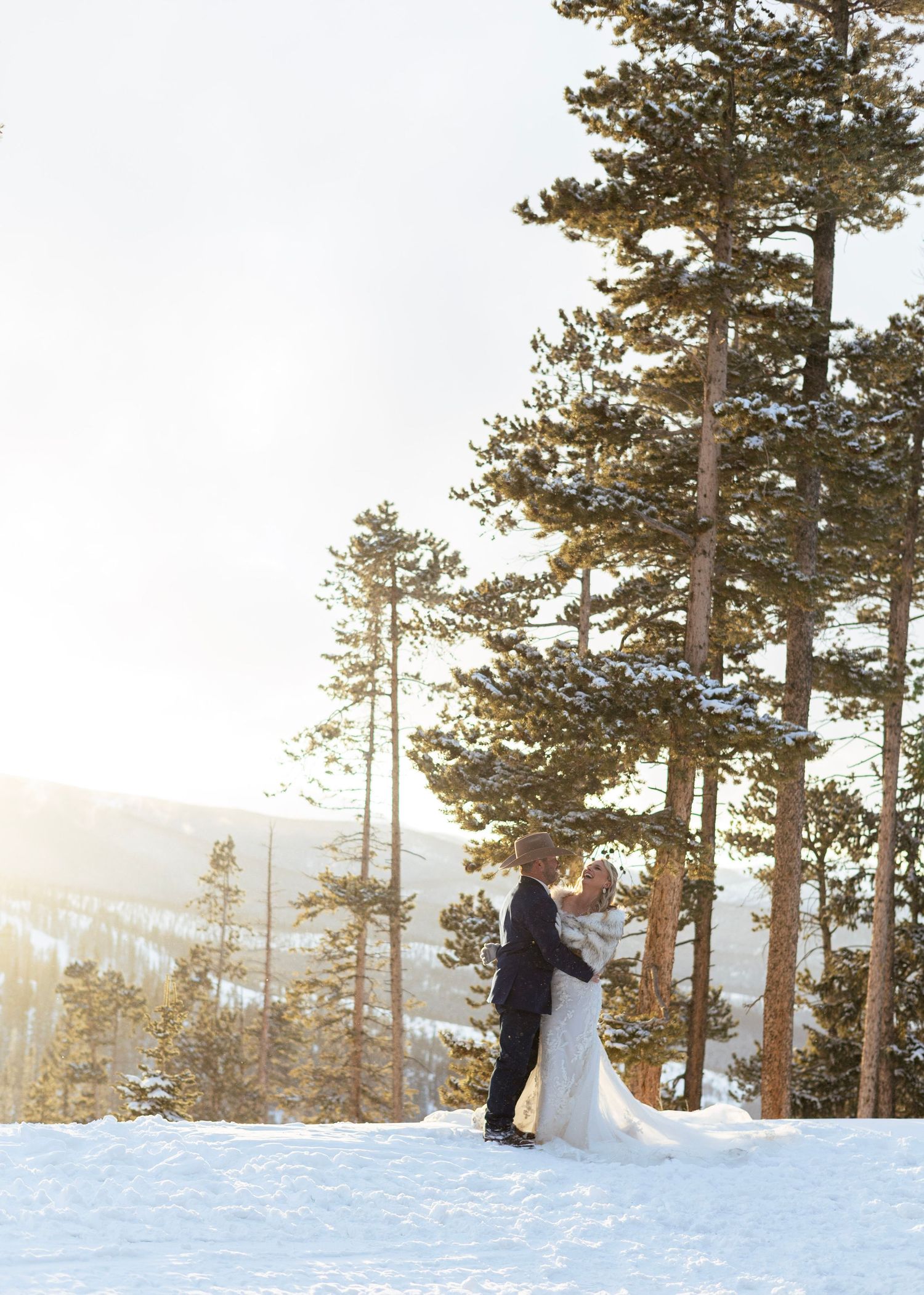 A wedding couple stands together on a snowy mountain overlook with tall pine trees at sunset.