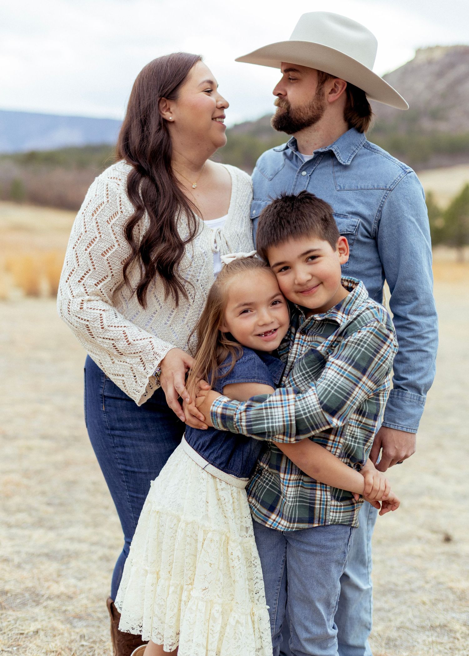 A happy family of four poses outdoors, parents in denim and cowboy hat, children hugging in a scenic field.