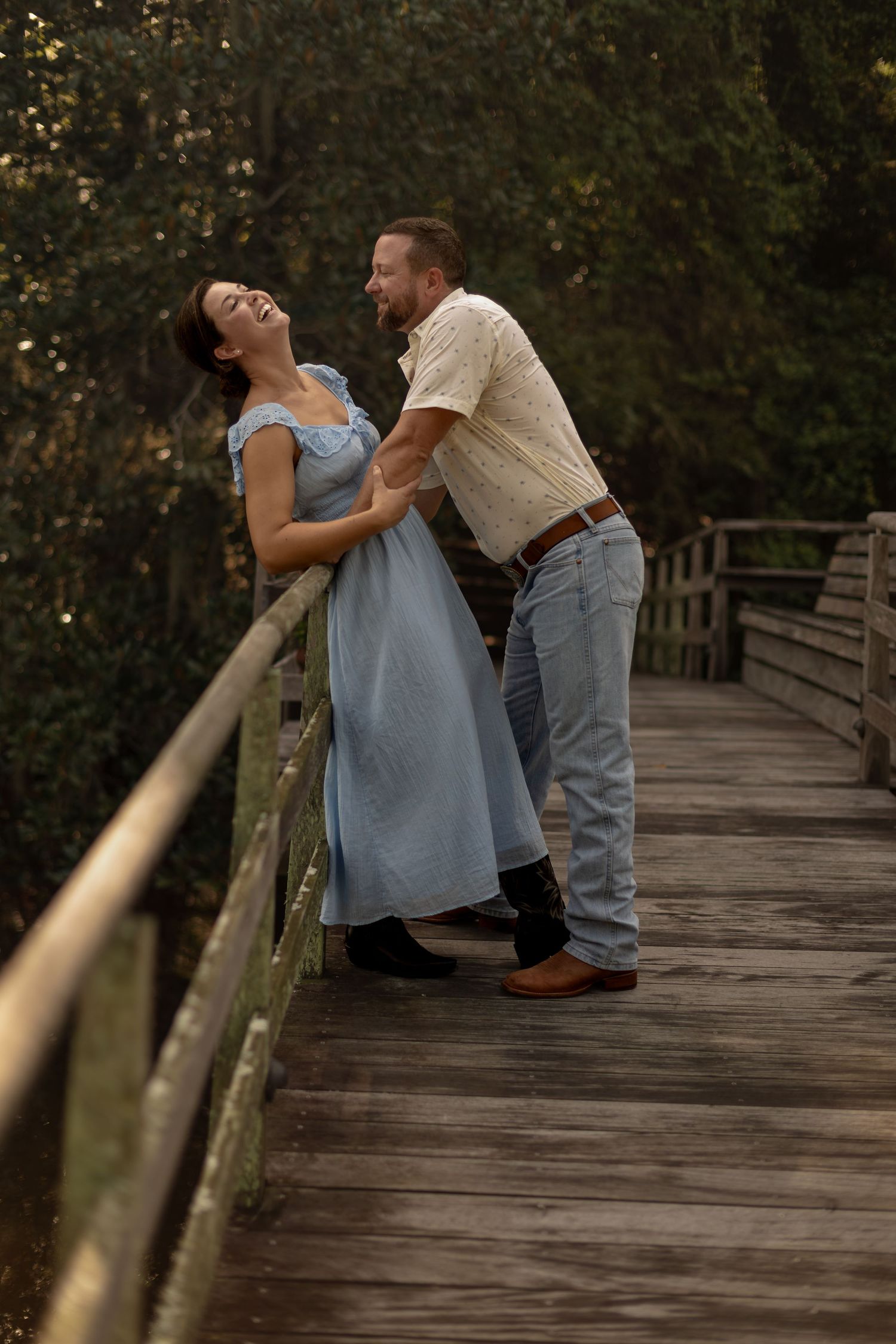 A joyful couple dances on a wooden bridge surrounded by lush greenery, she in a blue dress, he in jeans.