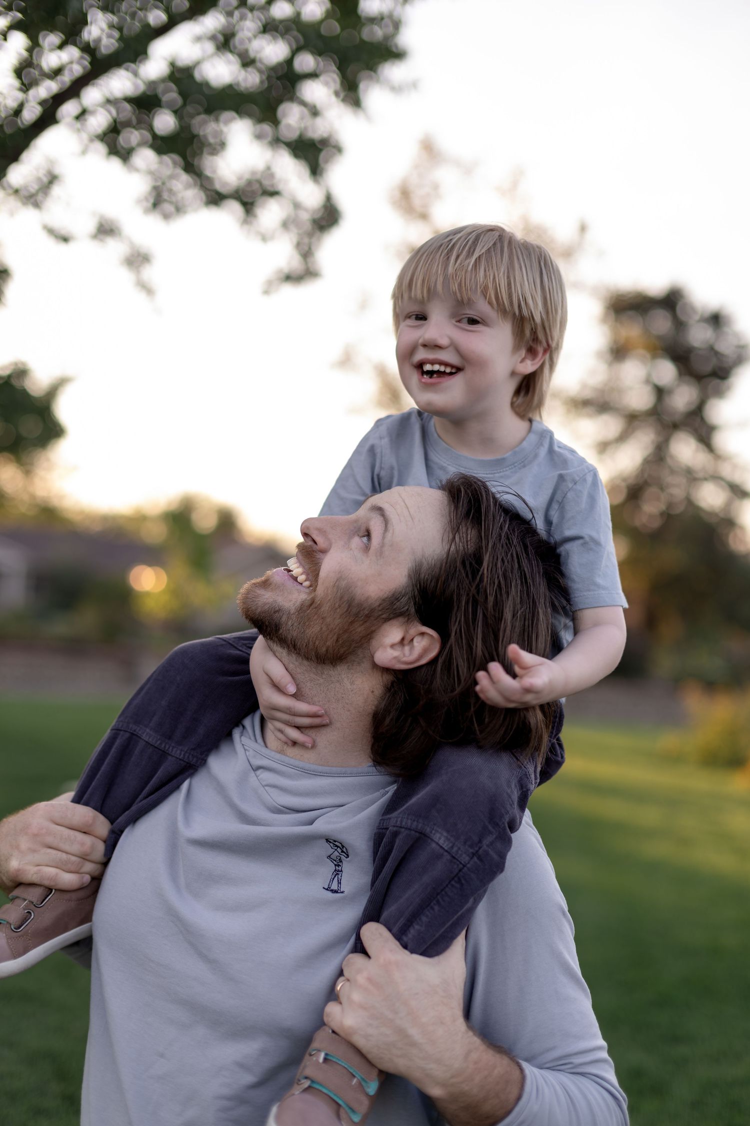 A laughing child sits on a bearded man's shoulders outdoors at sunset in a green park.