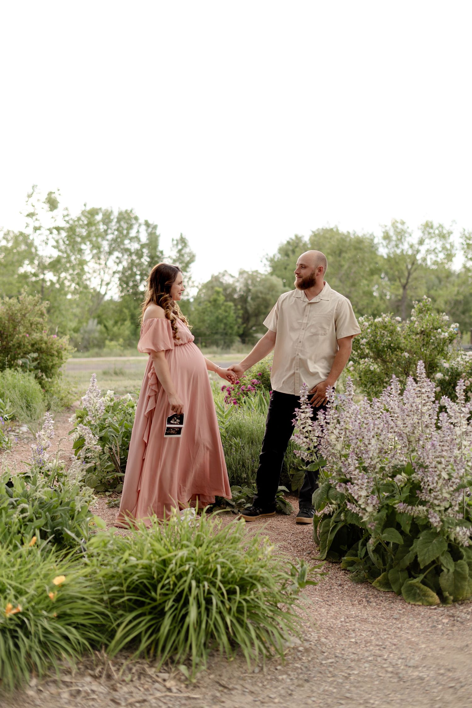 Expecting couple holding hands on a garden path, woman in blush maternity gown among purple wildflowers.