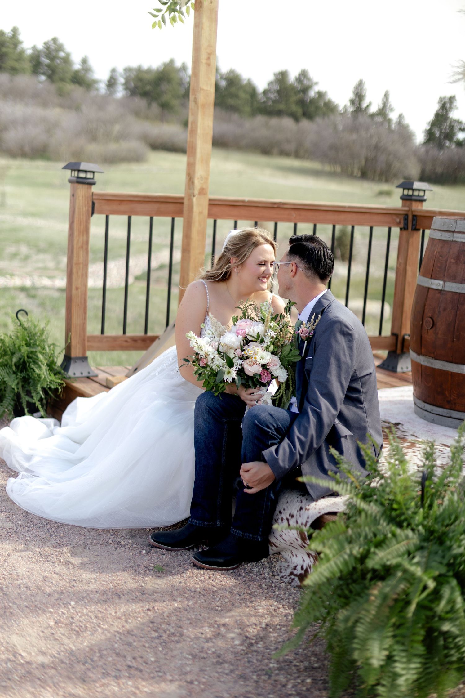 Bride and groom sitting together near a wooden arch, smiling and holding a floral bouquet outdoors.