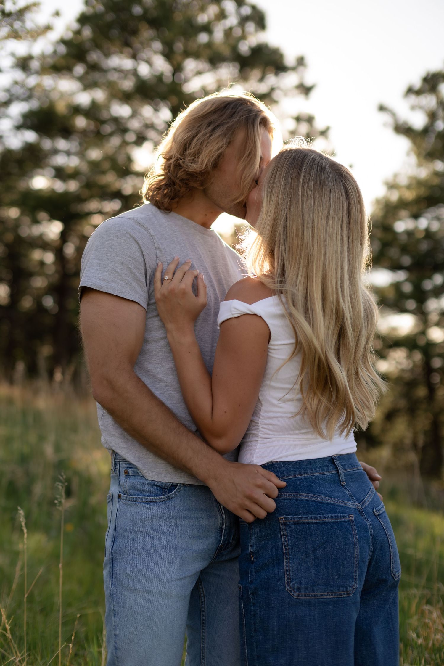 Romantic couple kissing outdoors in a grassy field during golden hour with pine trees behind them.