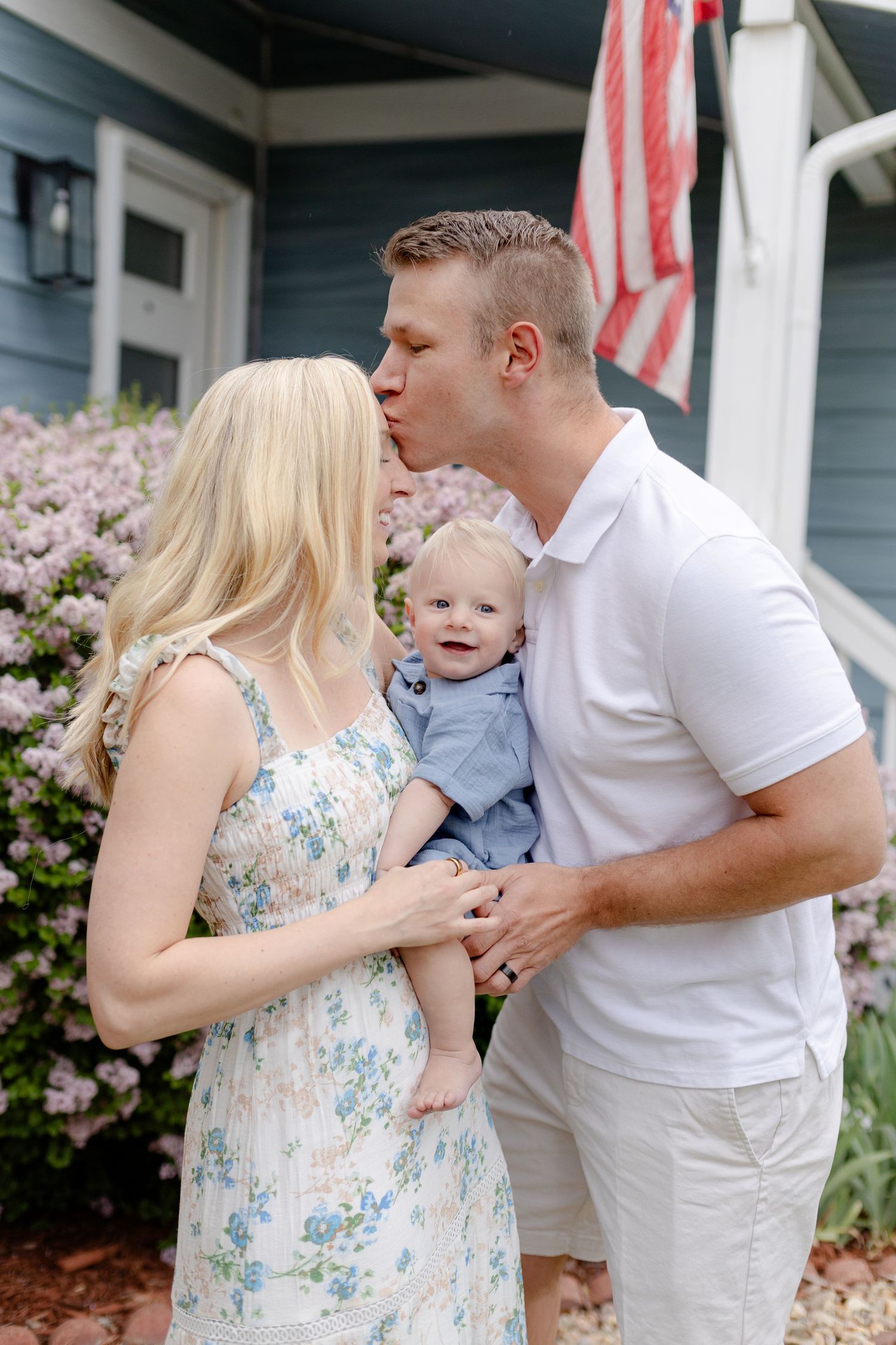 A couple kisses while holding their baby boy in front of a blue house with an American flag and pink flowers.